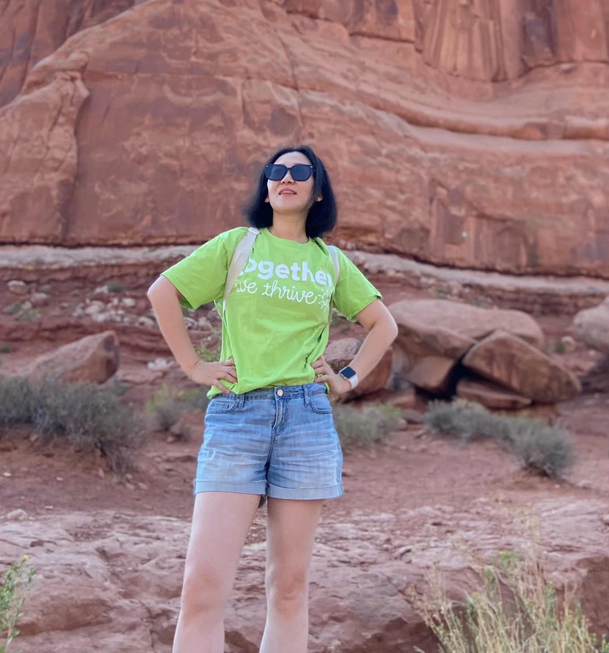 Woman wearing sunglasses and a bright green T-shirt standing outdoors in front of red rock formations with her hands on her hips.