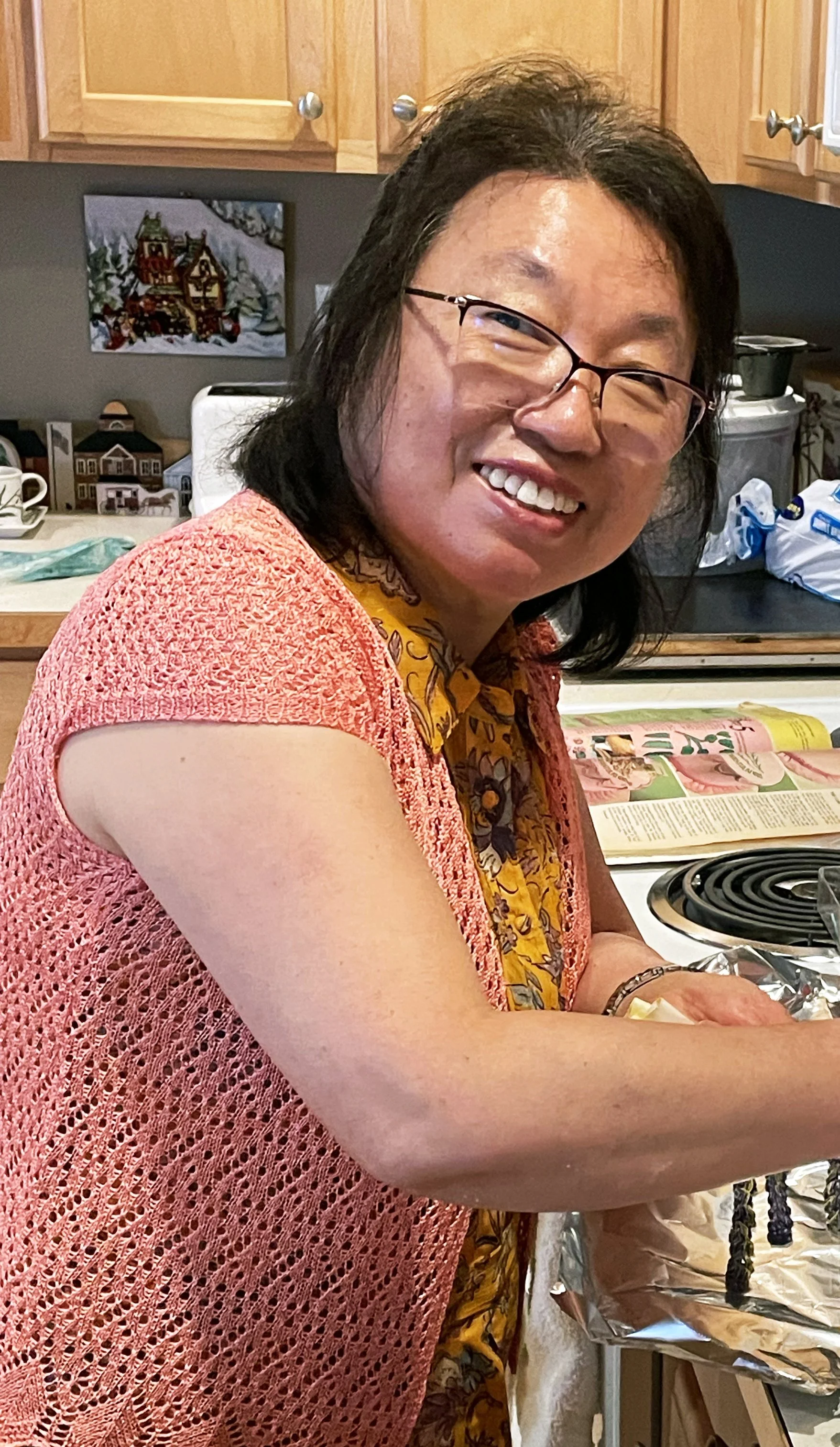 A woman with glasses smiling in a kitchen, wearing a pink crocheted top and a patterned yellow scarf, with kitchen cabinets, a stove, and holiday decorations in the background.
