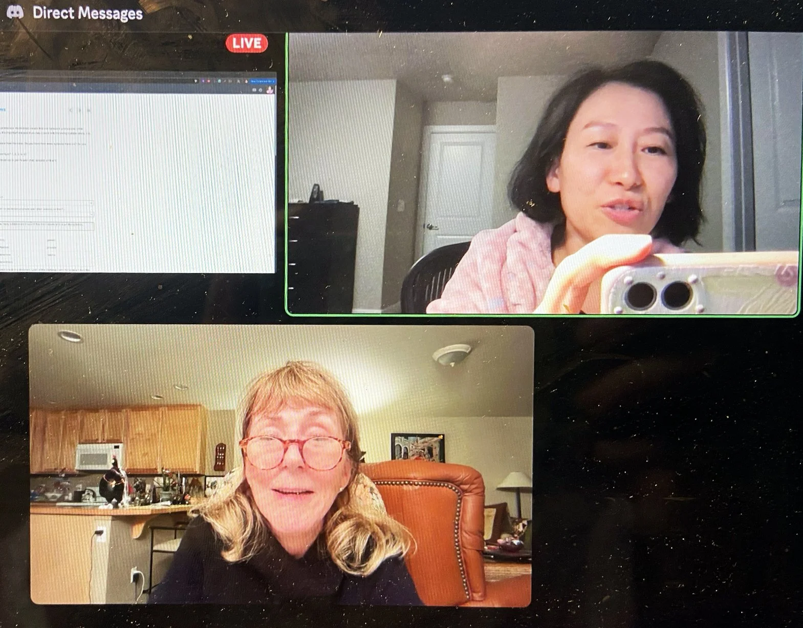A screenshot of a video call showing two women in separate locations. The woman in the top right corner has short dark hair, is wearing a pink hoodie, and is taking a selfie with her phone. The woman in the bottom left corner has long blond hair, glasses, and is smiling in a room with wooden furniture and kitchen items in the background.