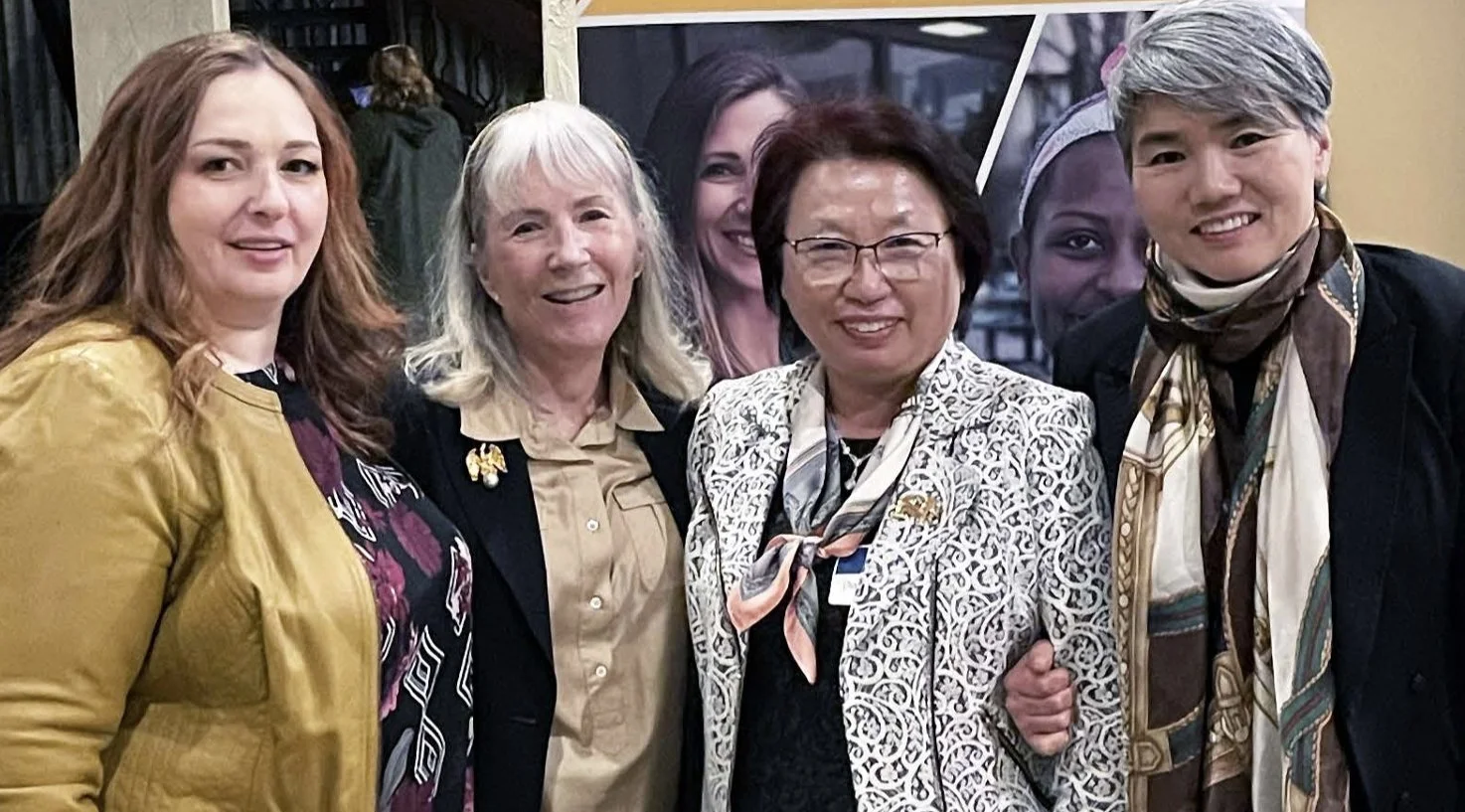 Group of five women standing together and smiling for a photo at an indoor event.