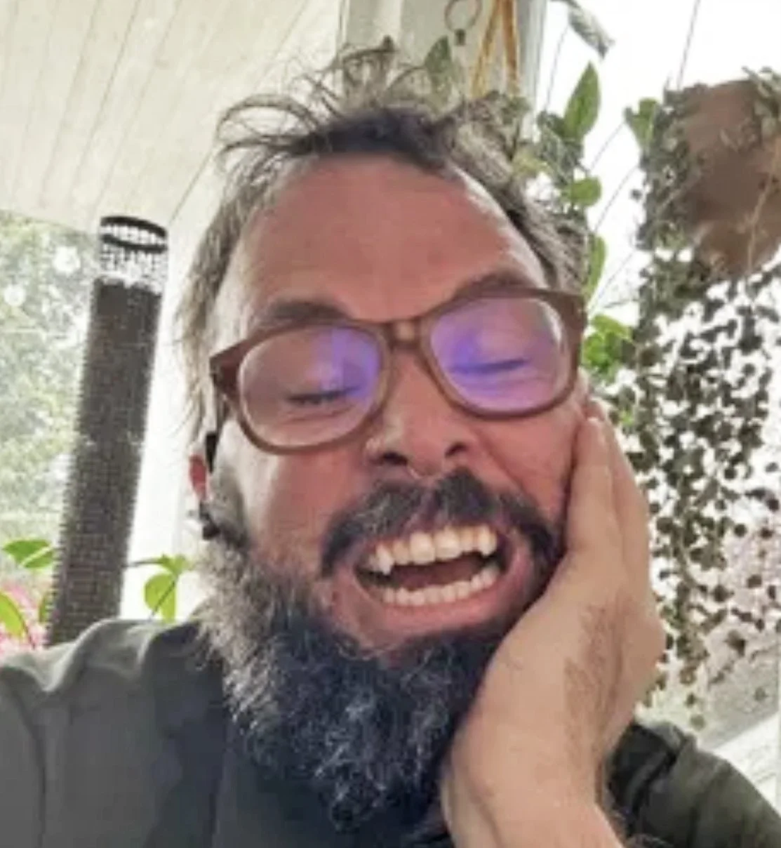 A man with glasses, a beard, and a big smile, resting his head on his hand indoors, with plants and a window in the background.