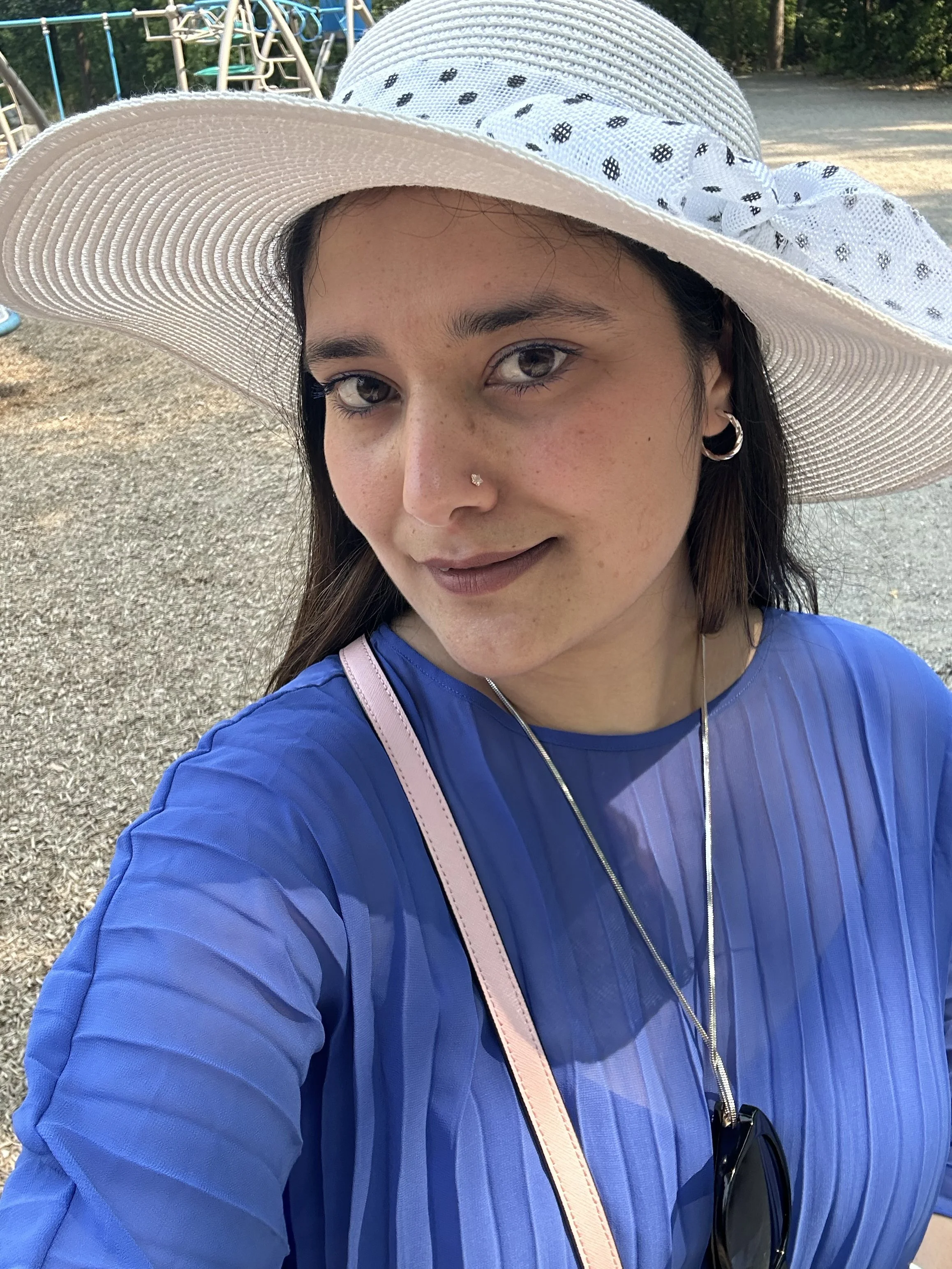 A woman wearing a large white sunhat with a dotted ribbon, a blue jacket, and earrings, standing outdoors near a playground with trees in the background.