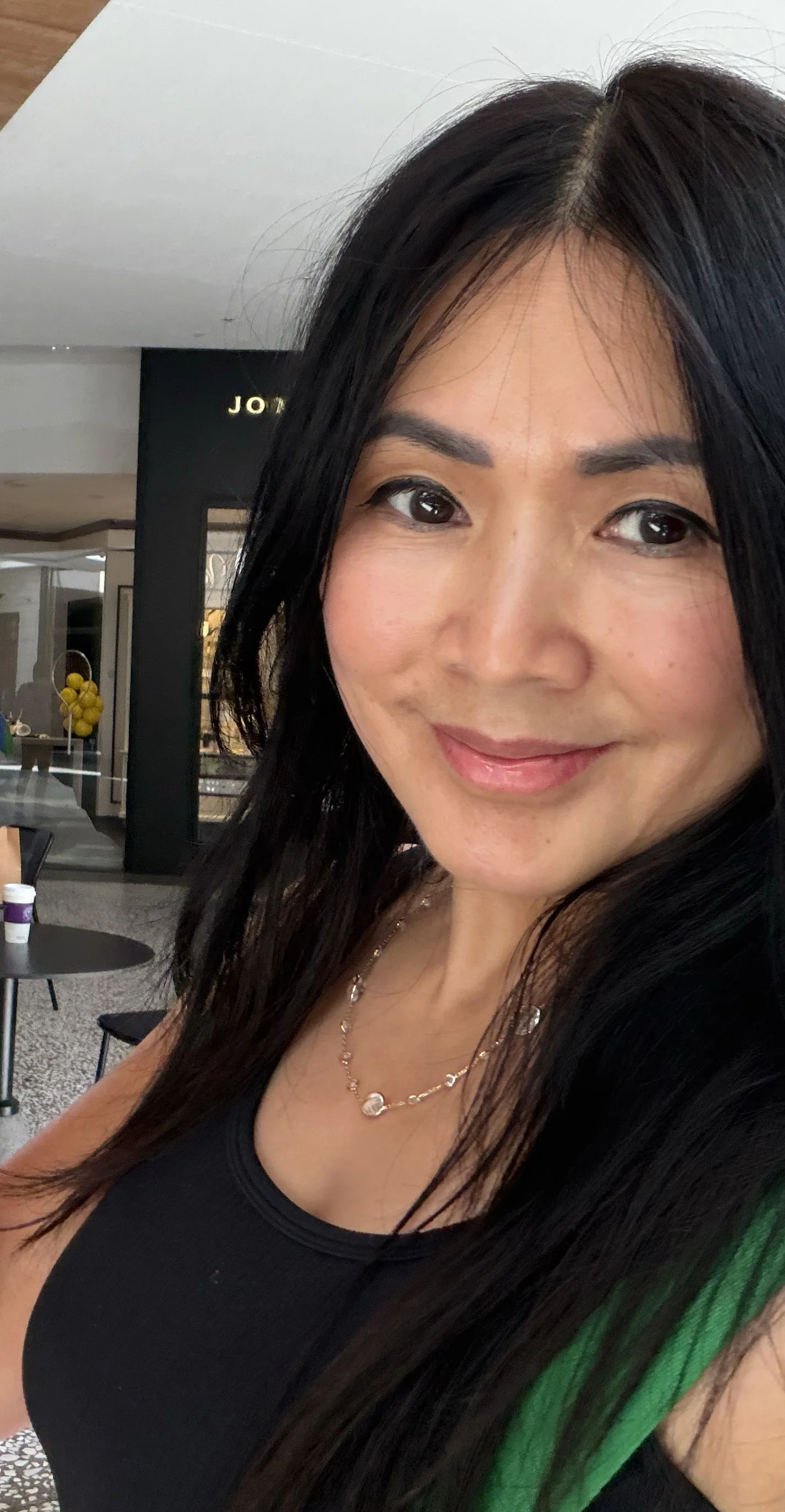 Close-up of a woman with long black hair, smiling, wearing a black top and a necklace, inside a modern mall or shopping center.