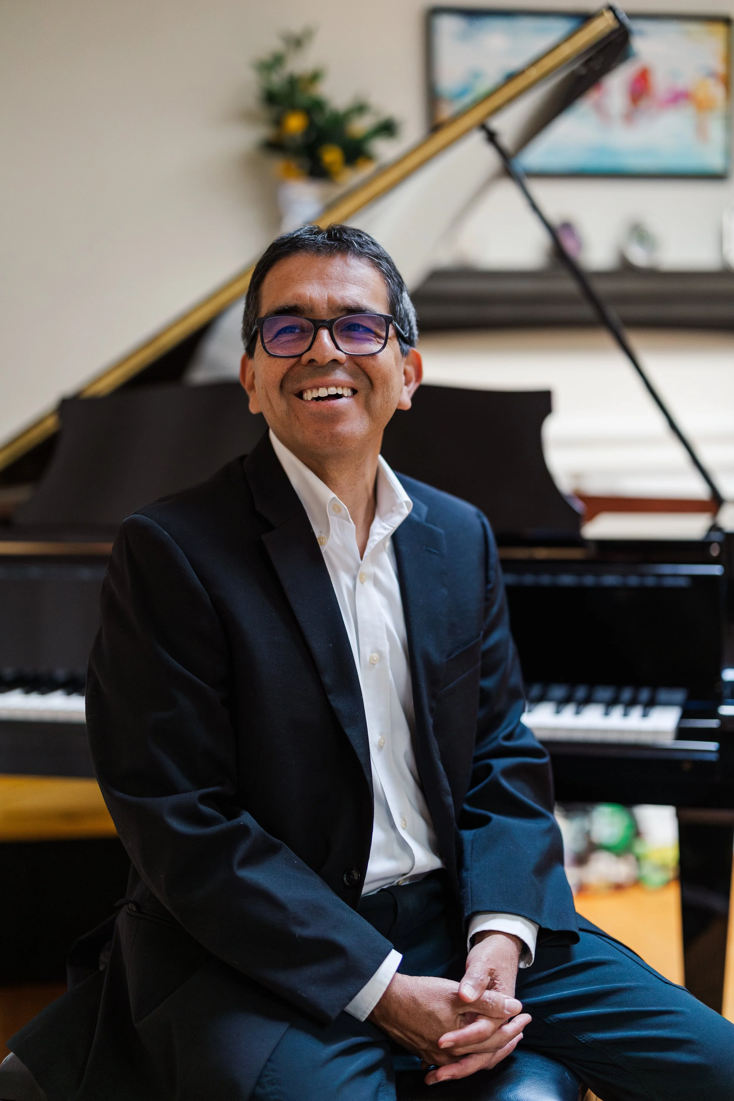 A man wearing glasses, in a suit, smiling, seated in front of a grand piano inside a room decorated with a flower arrangement and framed artwork on the wall.