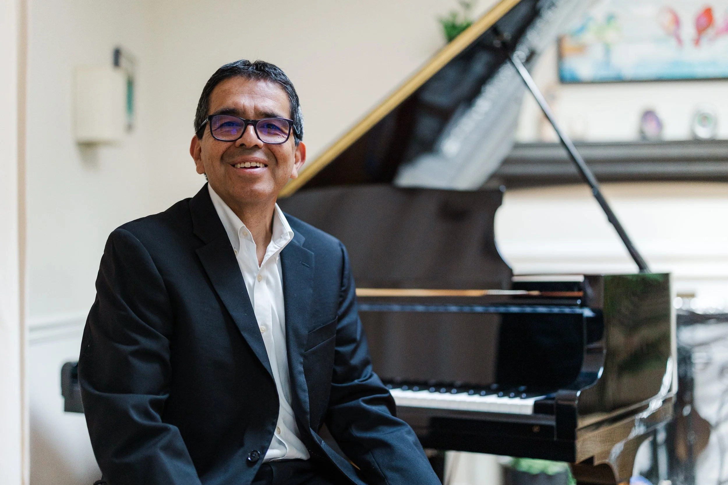Man in a black suit and white shirt sitting at a grand piano, smiling, in a bright room with a window and wall decorations.