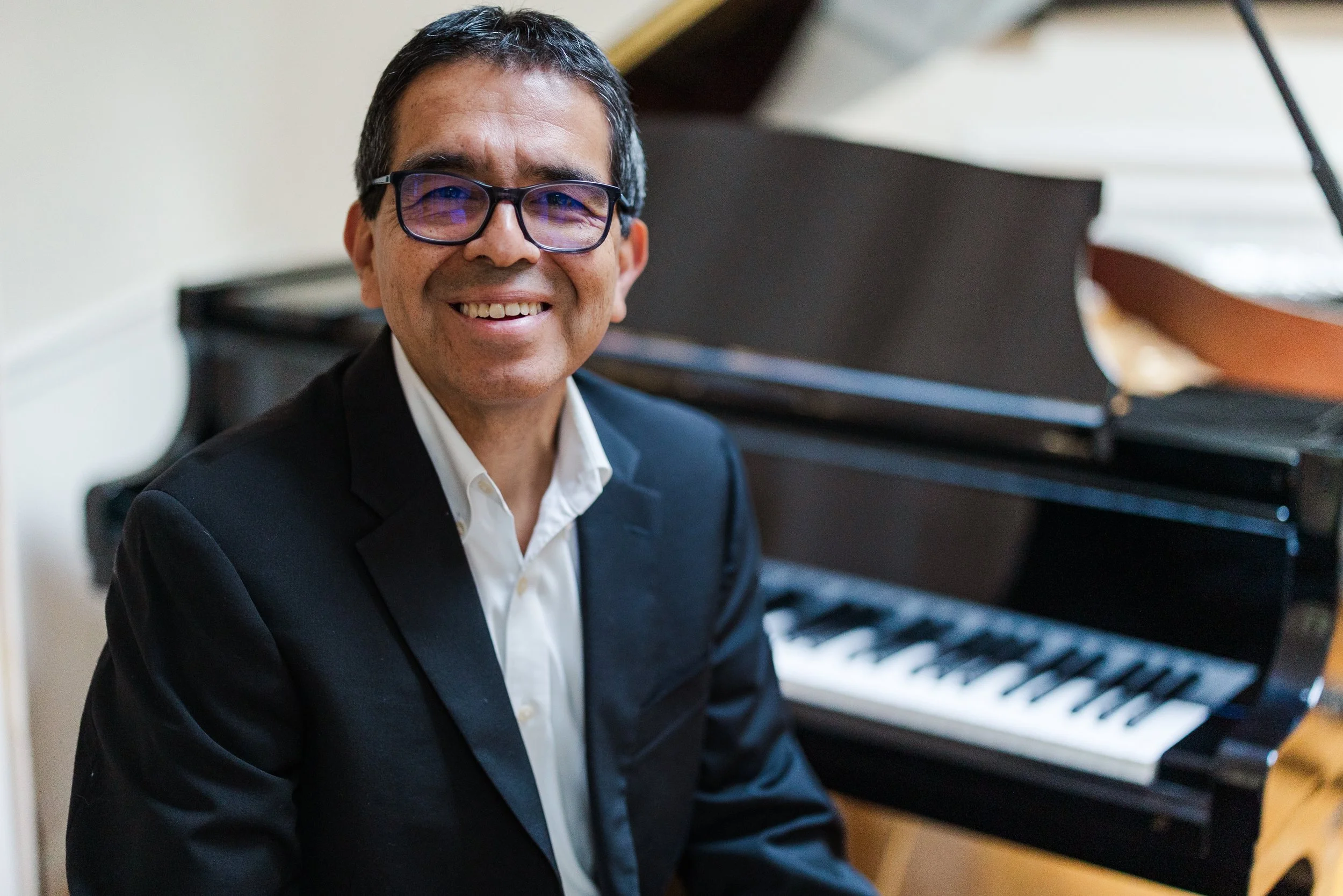A smiling man wearing glasses in a black suit and white shirt, sitting at a piano with black and white keys, in a room with white walls.