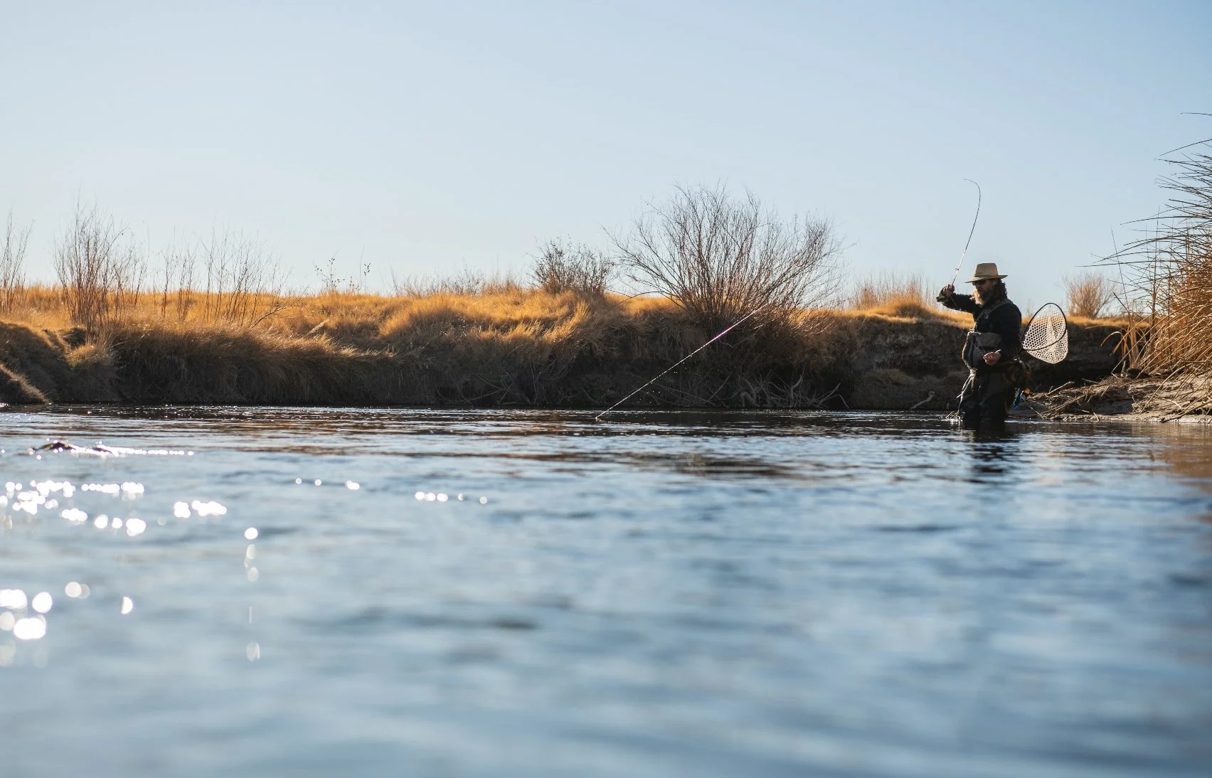 A person fishing in a river with dry grass and bushes along the riverbank, under a clear sky.