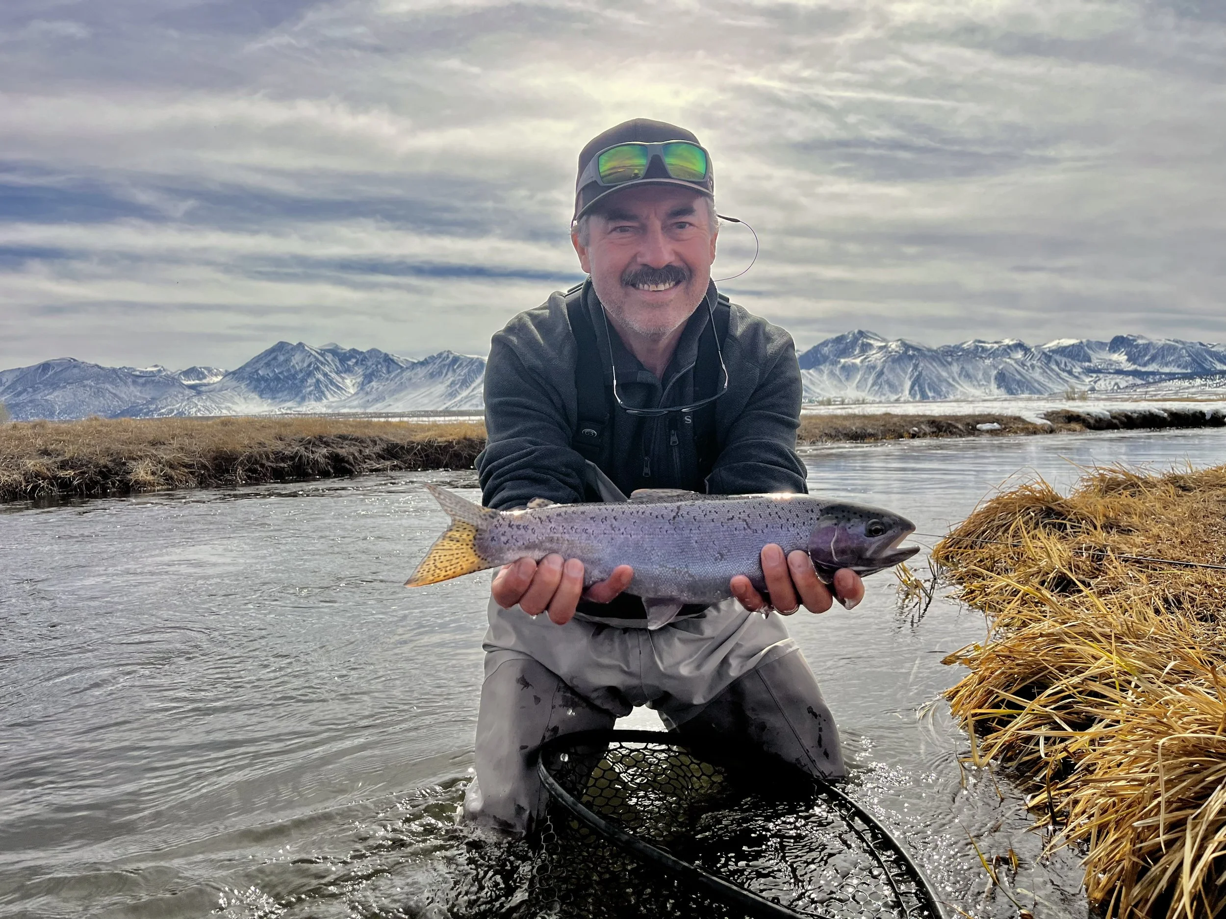 A man with a big smile, dressed in outdoor gear, kneeling in a river, holding a large fish with mountains in the background.