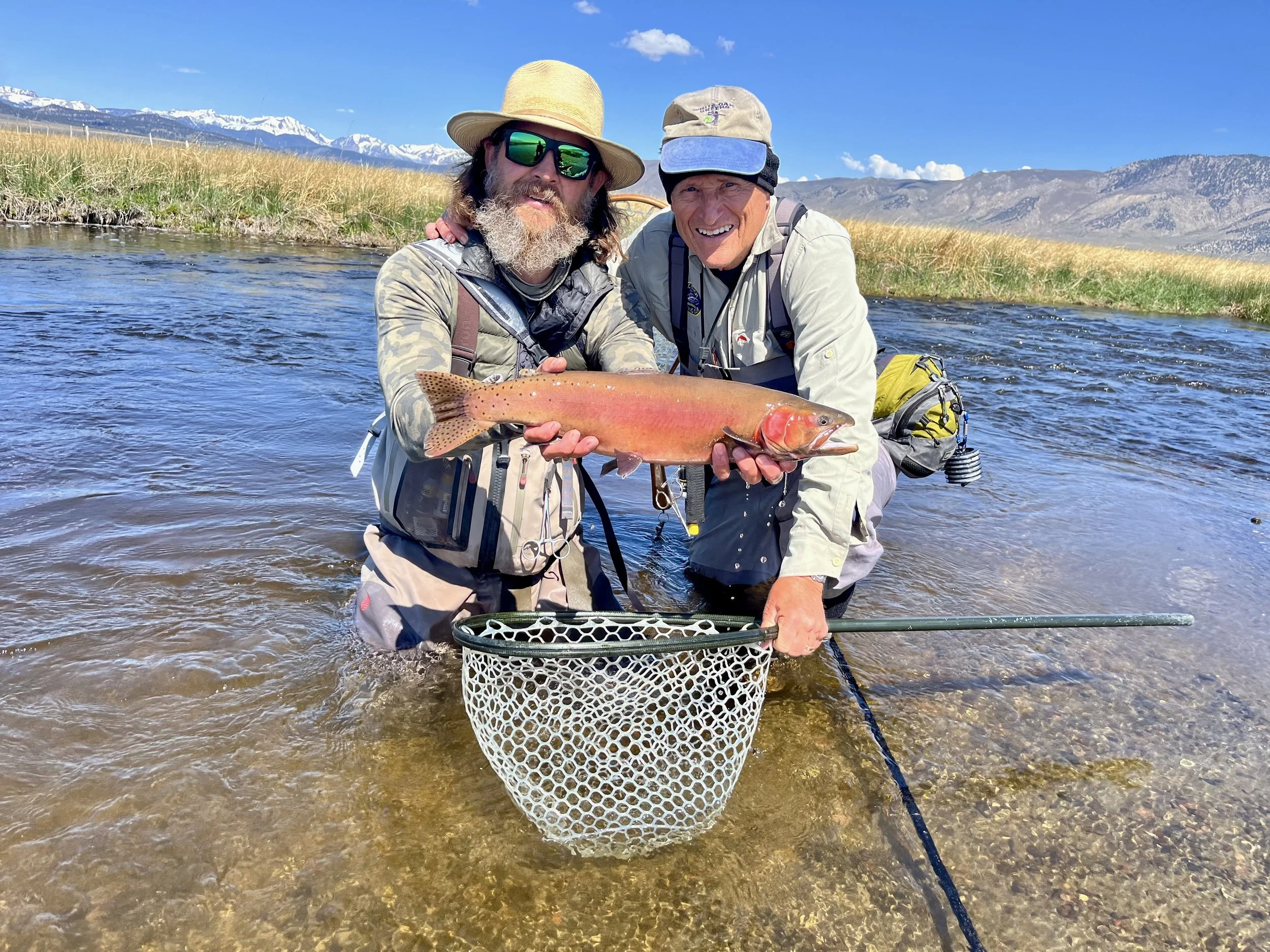 Two men standing in a shallow river, holding a large fish, with mountains and a grassy landscape in the background on a clear day.
