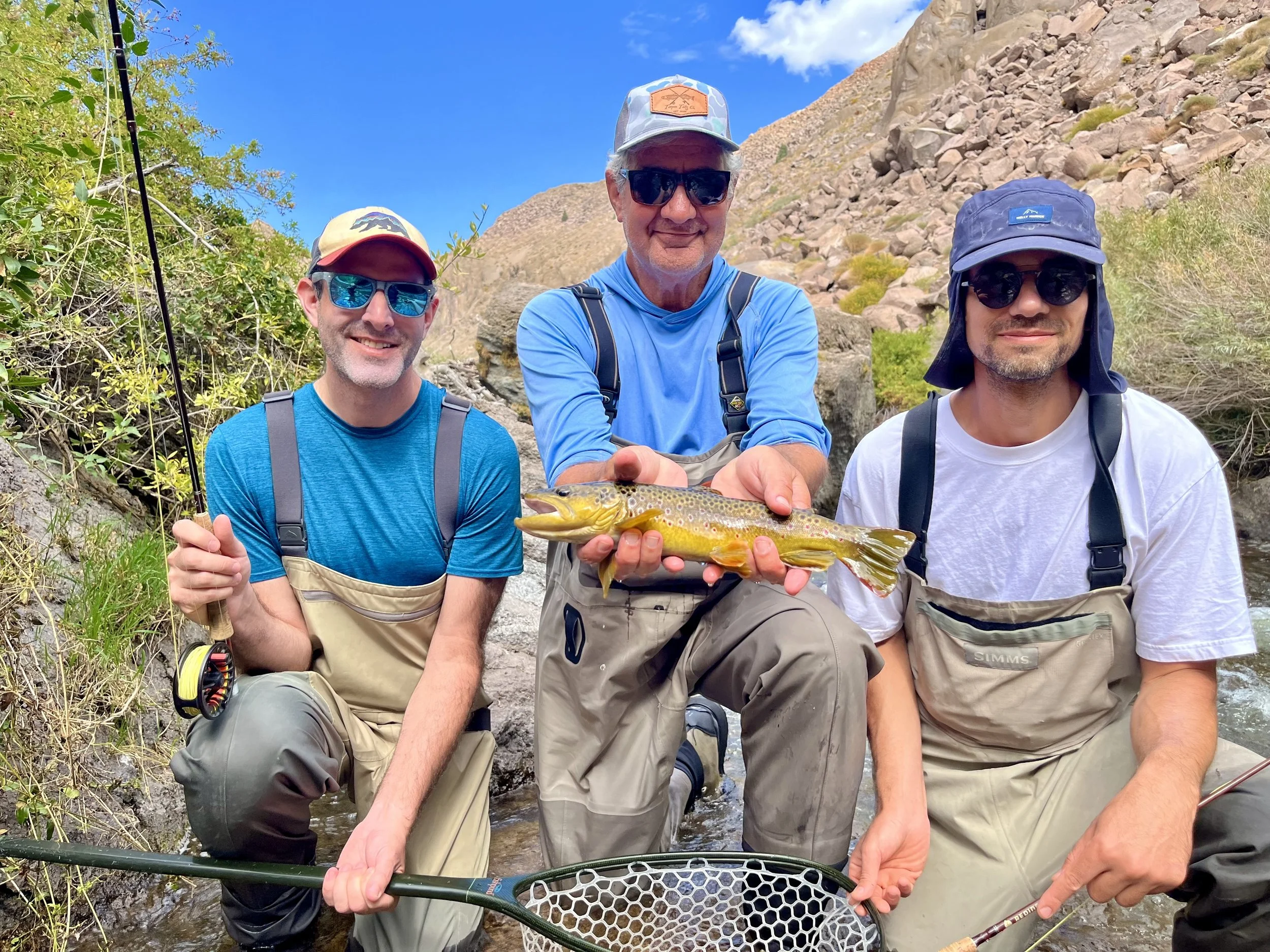 Three men on a rocky riverbank, wearing fishing gear, posing with a fish they caught during a fishing trip.