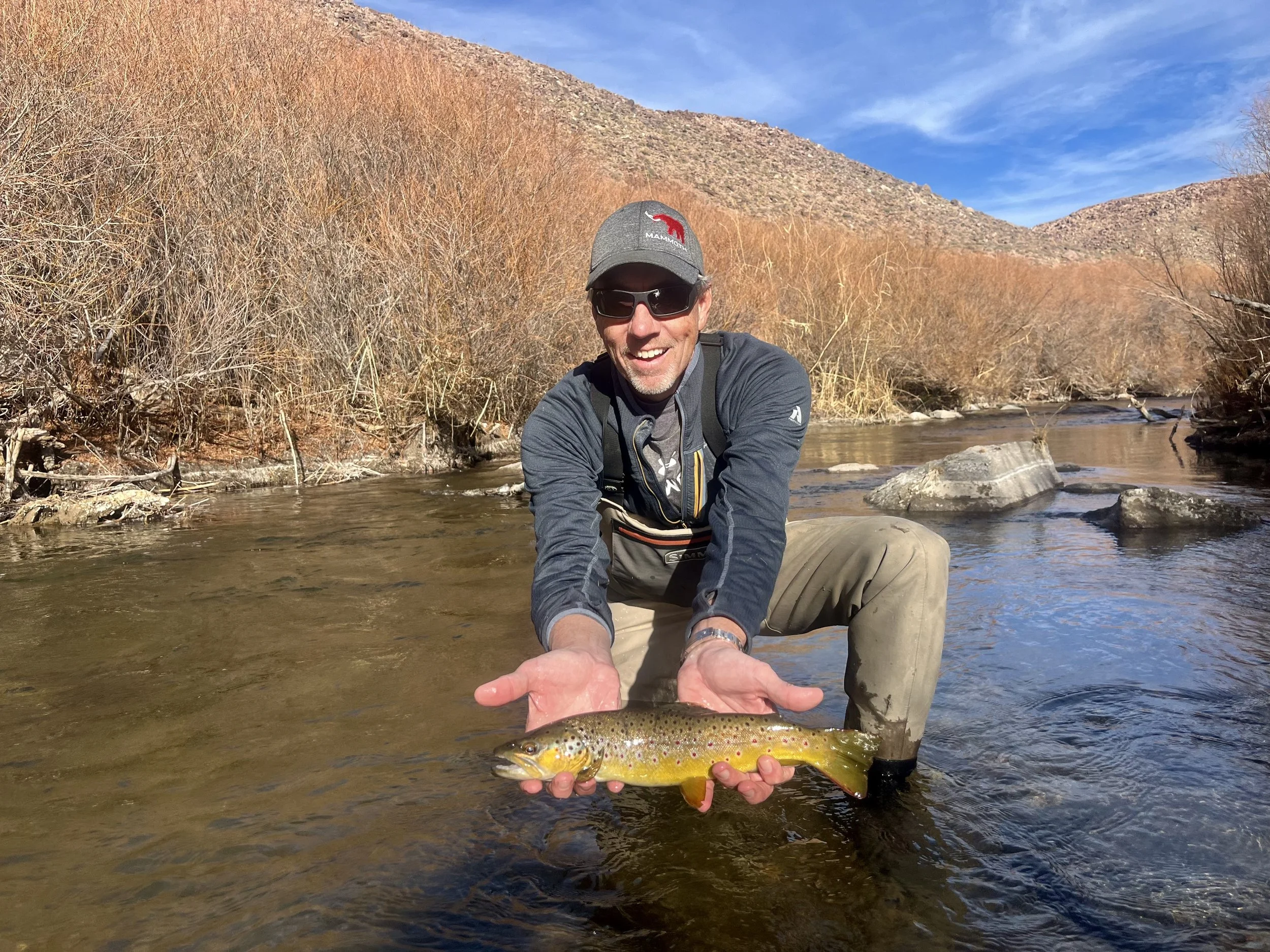 Man wearing sunglasses, a gray cap, and outdoor clothing kneeling in a shallow river, holding a small rainbow trout with yellow and pink spots, surrounded by bare trees and mountains in the background.