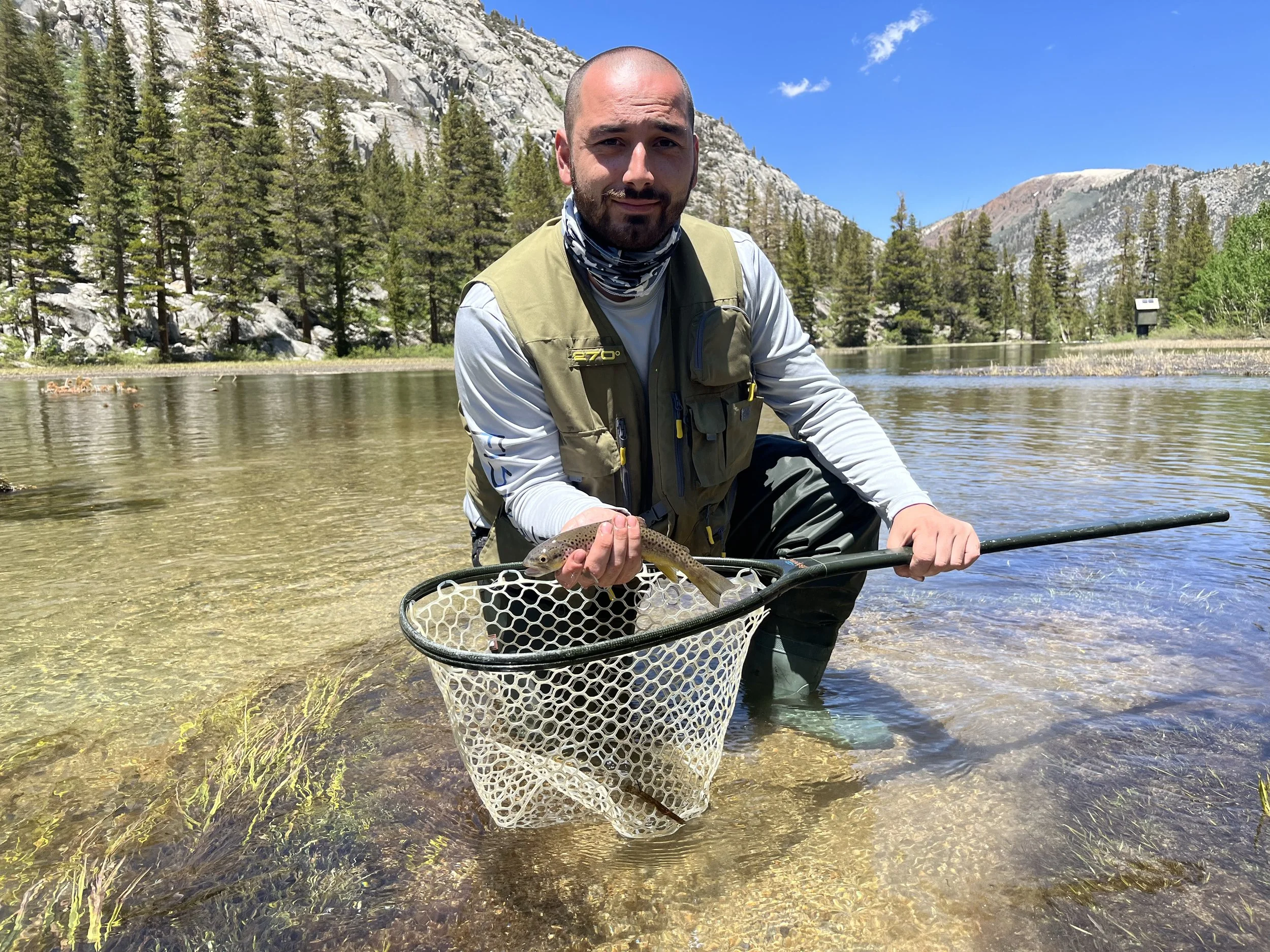Man kneeling in shallow water holding a fish with a fishing net in a scenic outdoor setting with trees and mountains.