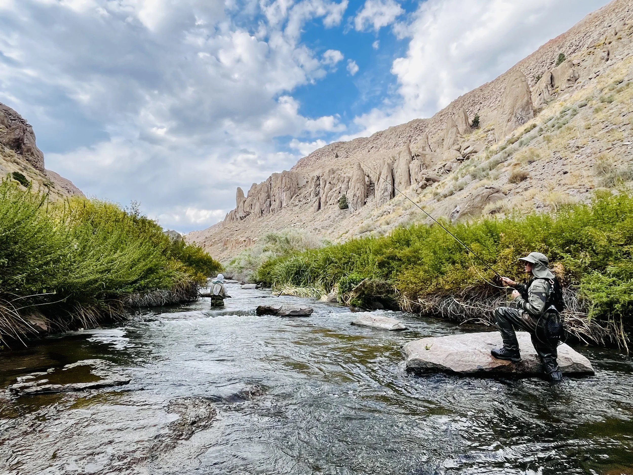 Two people fly fishing in a river surrounded by mountains and green bushes under a partly cloudy sky.