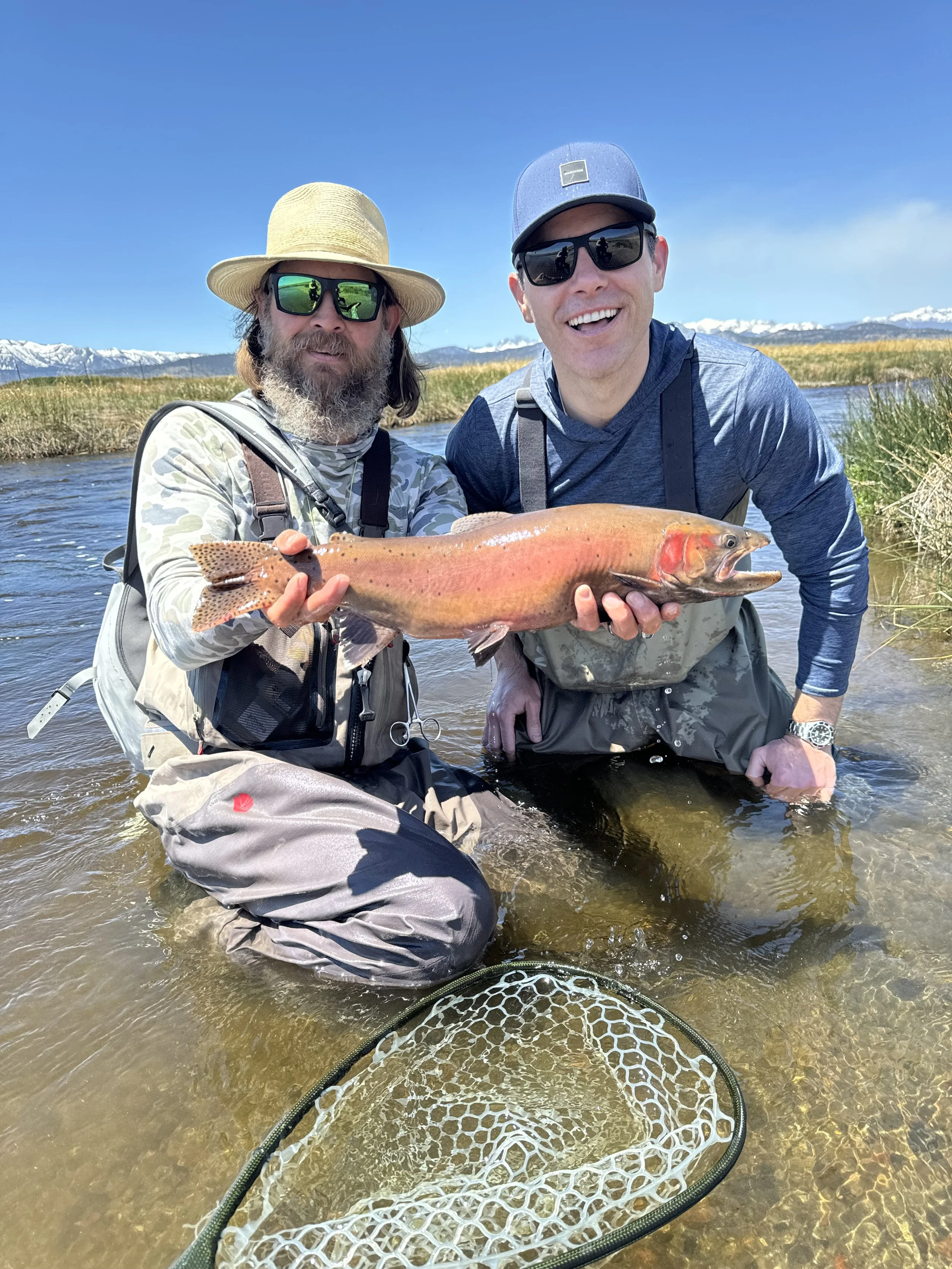 Two men standing in a river, holding a large fish, with mountains and clear blue sky in the background.