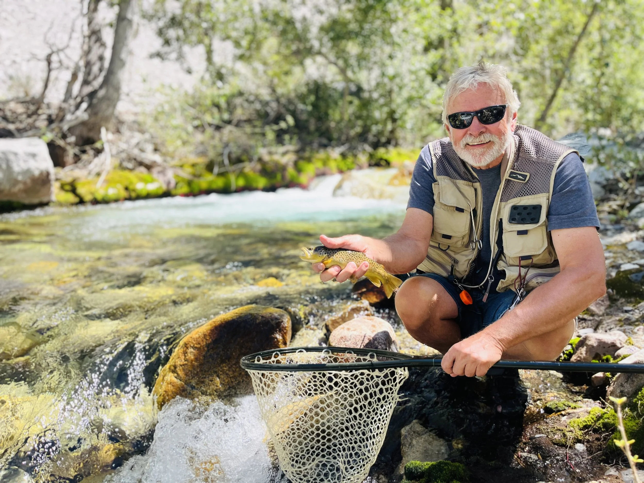 Senior man with white hair and beard, wearing sunglasses, kneeling in a creek holding a small fish, with a fishing net and rocks around him, surrounded by lush green trees.
