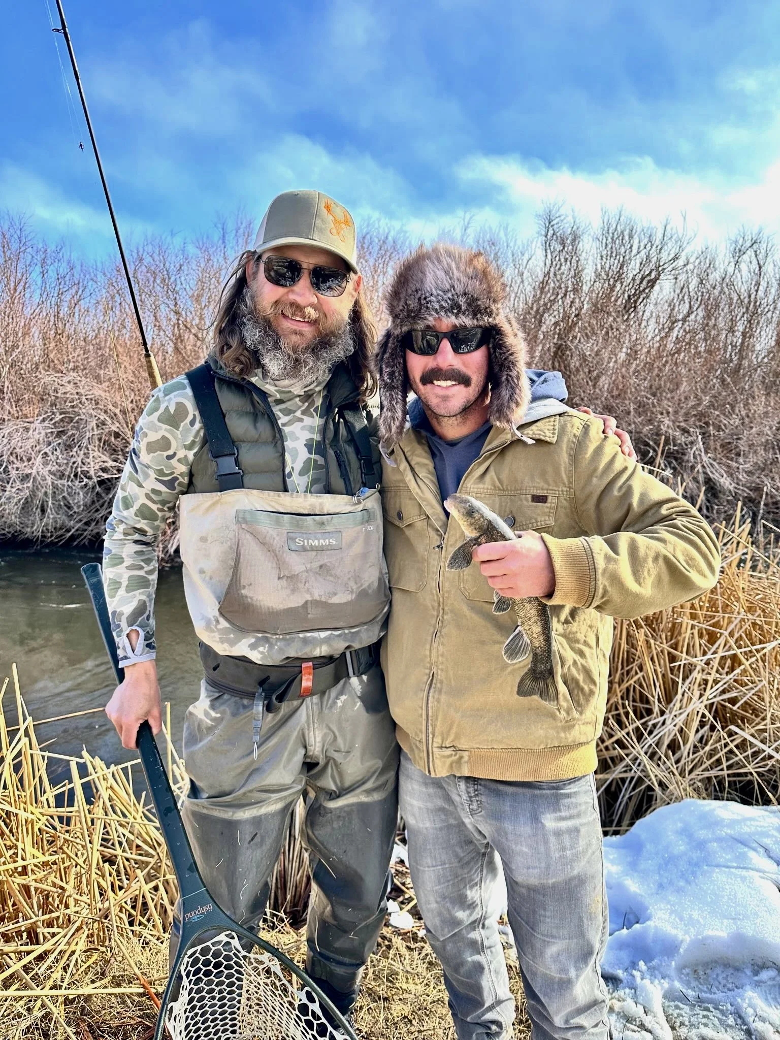 Two men smiling by a riverbank, one holding a fish, on a sunny winter day with snow on the ground and bushes in the background.