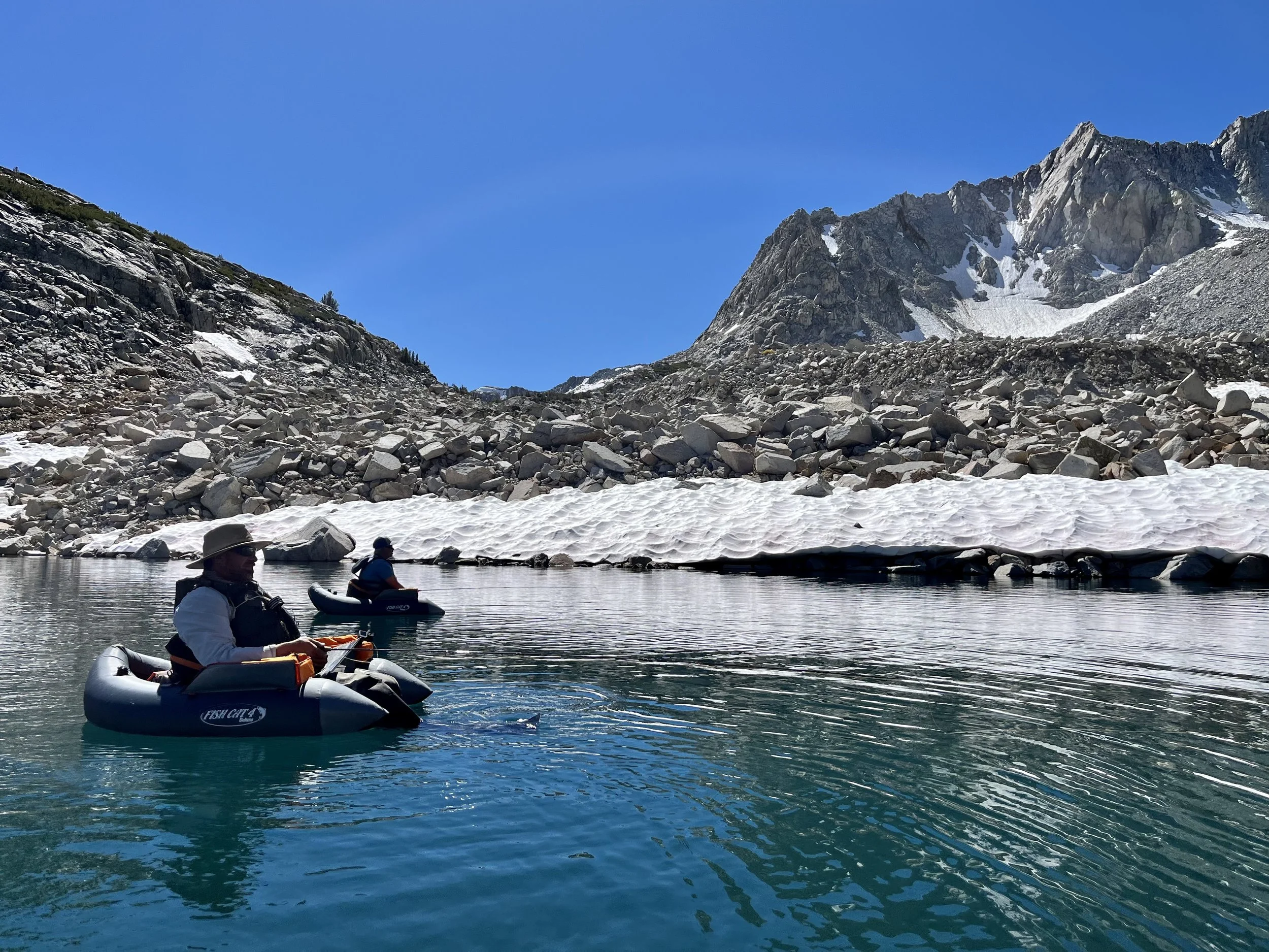 Two people in inflatable boats floating on a mountain lake surrounded by snow and rocky terrain, with mountain peaks in the background under a clear blue sky.
