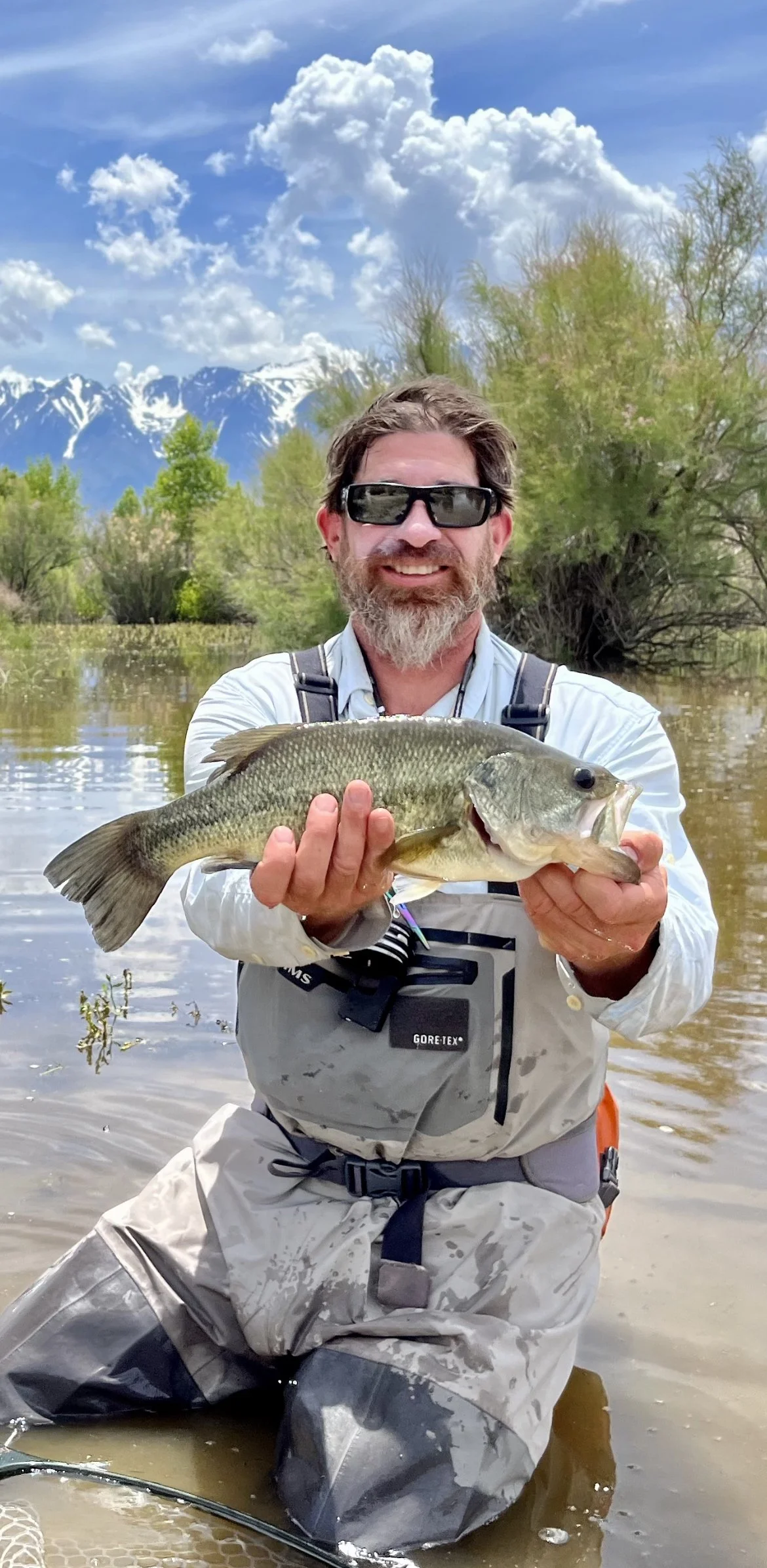 A man wearing sunglasses, a light-colored fishing outfit, and waders kneeling in shallow water, holding a large bass fish he caught during a fishing trip. Backdrop includes a river, green trees, snow-capped mountains, and a partly cloudy sky.