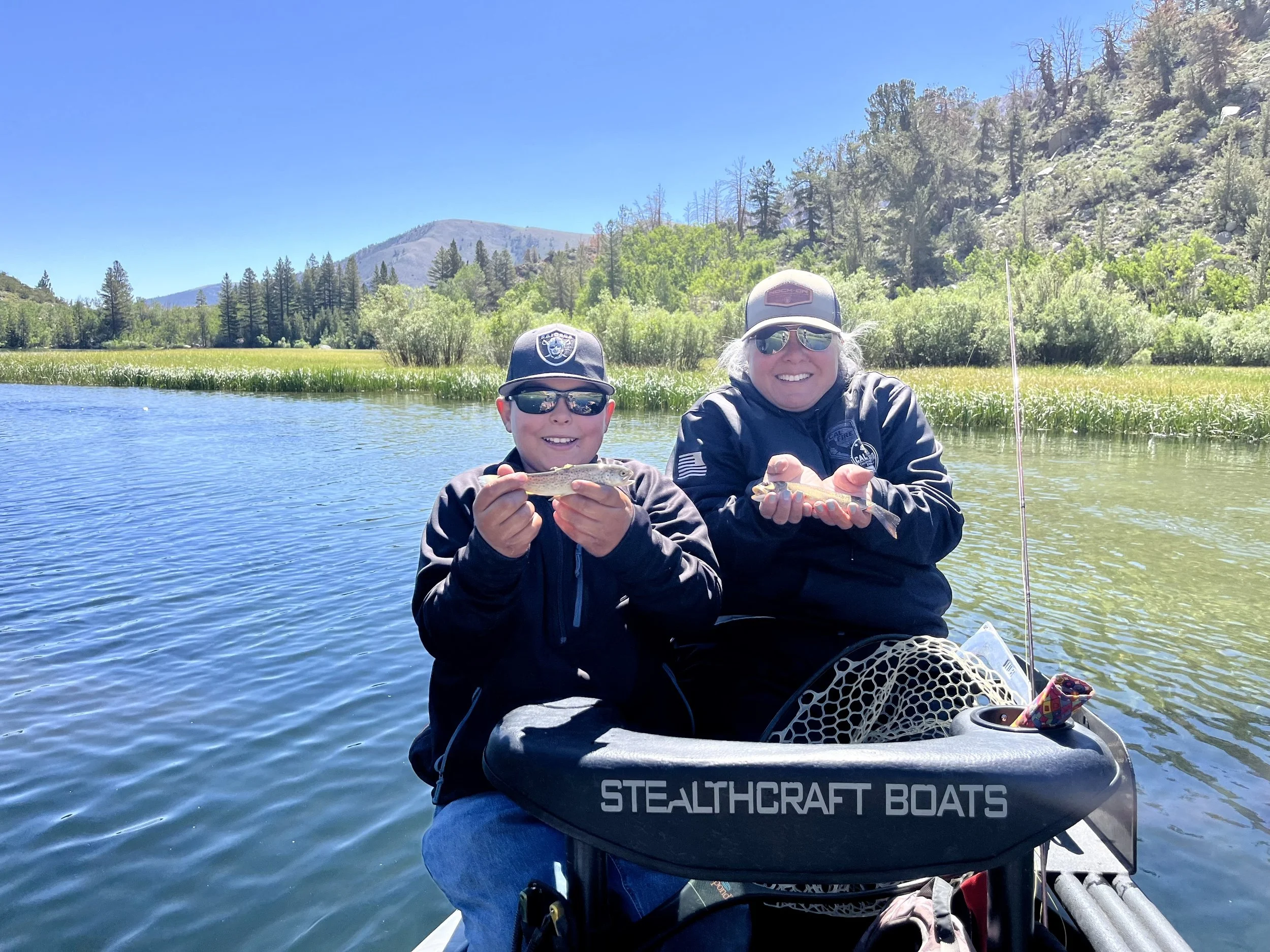 Two smiling people, a child and an adult, sitting in a small boat on a lake, holding small fish they caught, with a scenic backdrop of trees, grass, and mountains on a sunny day.