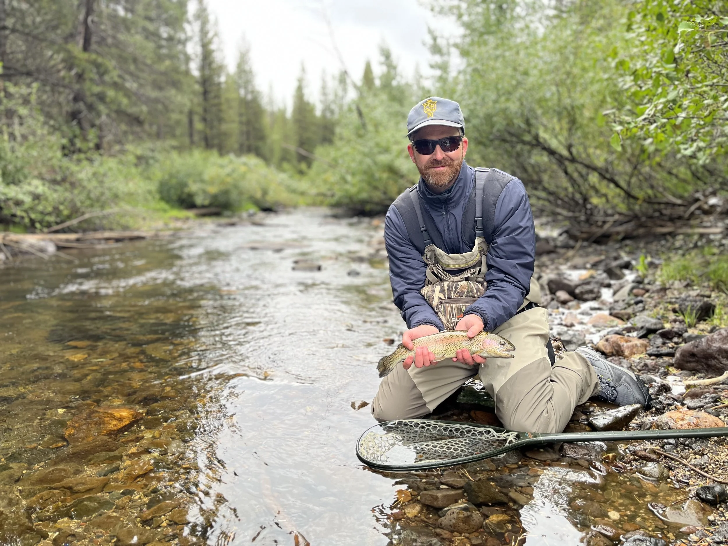 A man kneeling by a riverbank holding a caught fish, wearing sunglasses, a cap, a navy jacket, and khaki pants, with a fishing net at his side amid a forested landscape.