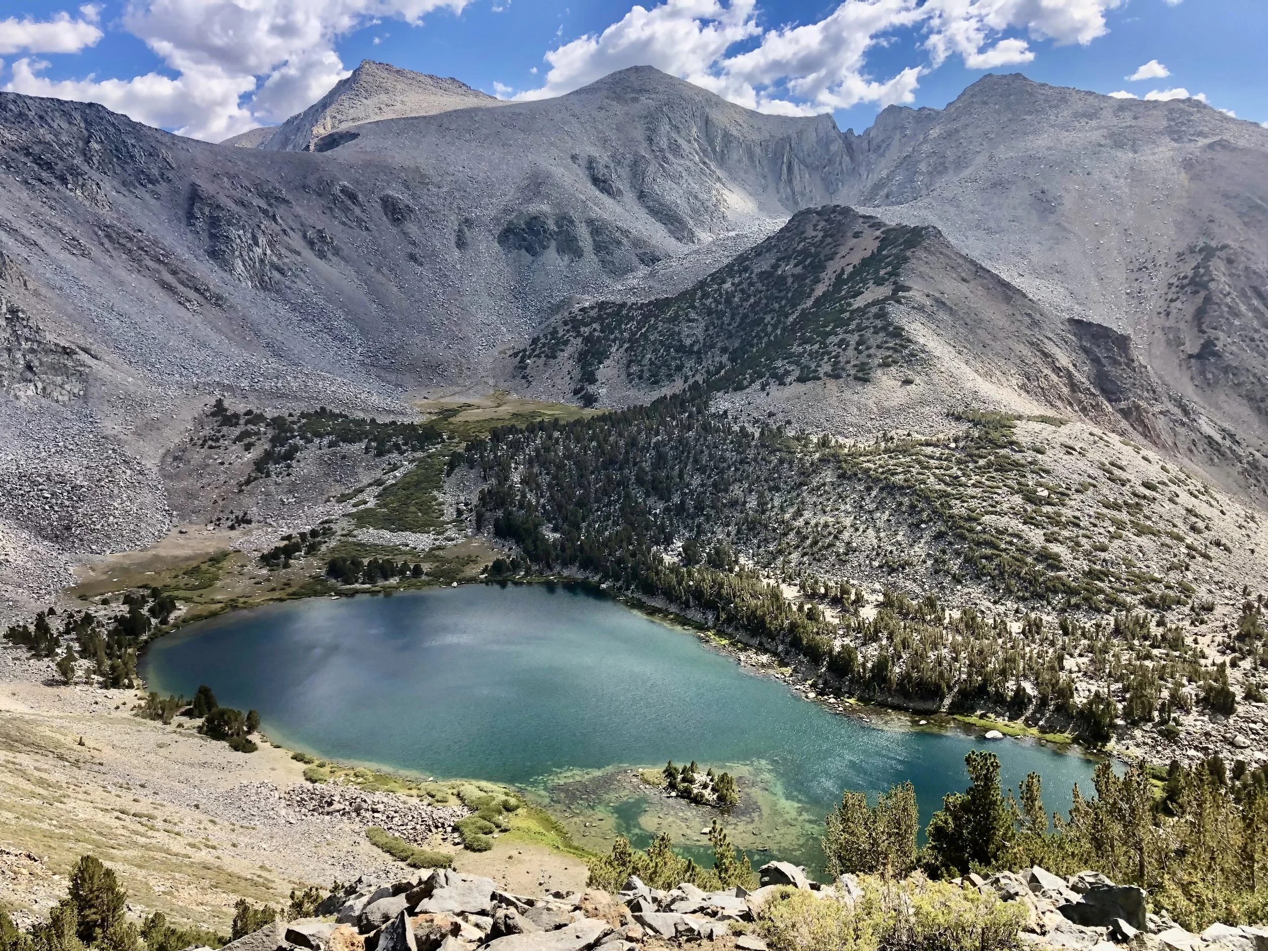 A high-altitude mountain landscape featuring a small, round lake surrounded by rocky slopes and sparse greenery, with tall mountains in the background under a partly cloudy sky.