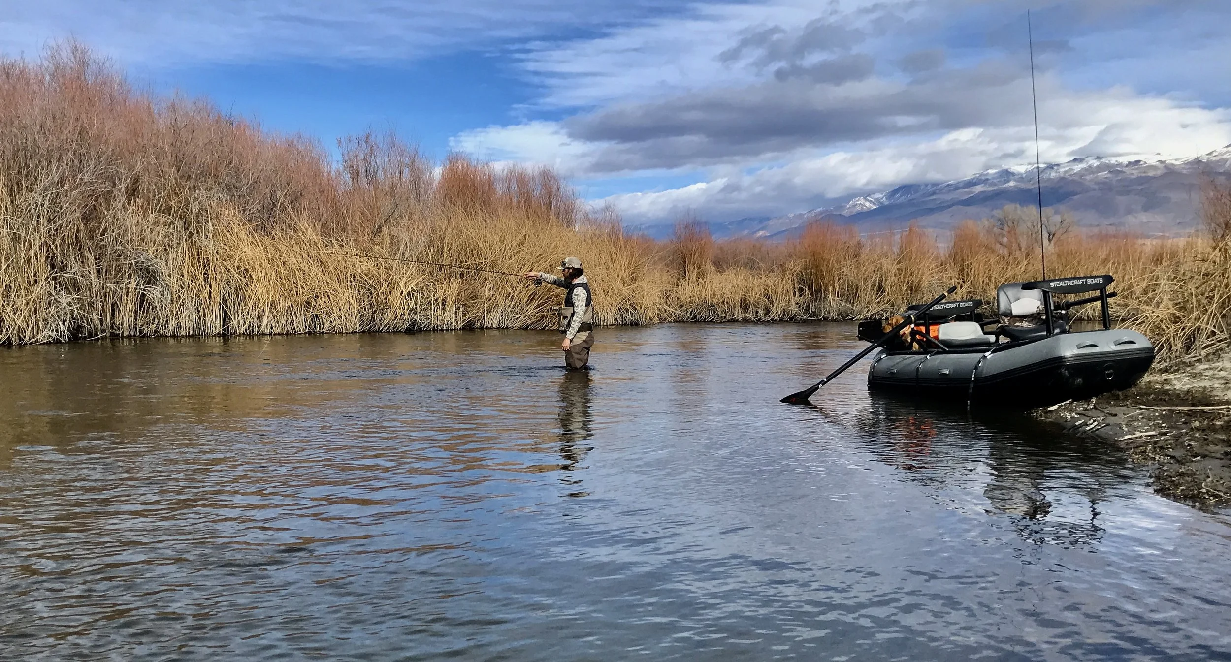 Person fishing in a river with a boat on the shore, dry bushes, mountain range in the background, and cloudy sky.