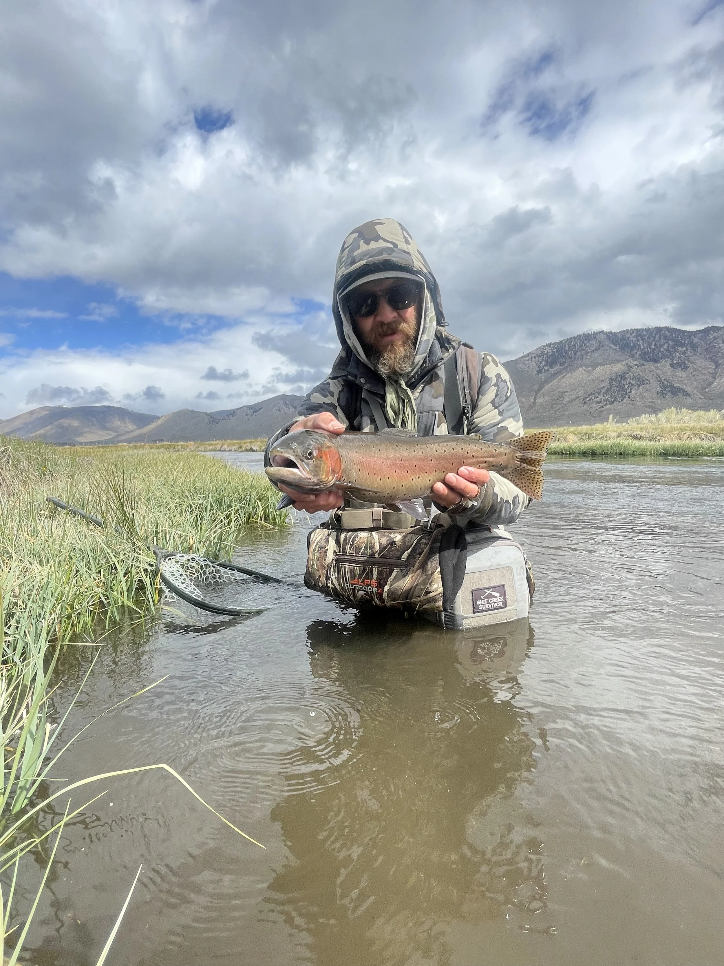 Man in camouflage clothing and sunglasses holding a fish in a shallow river with mountains and cloudy sky in the background.