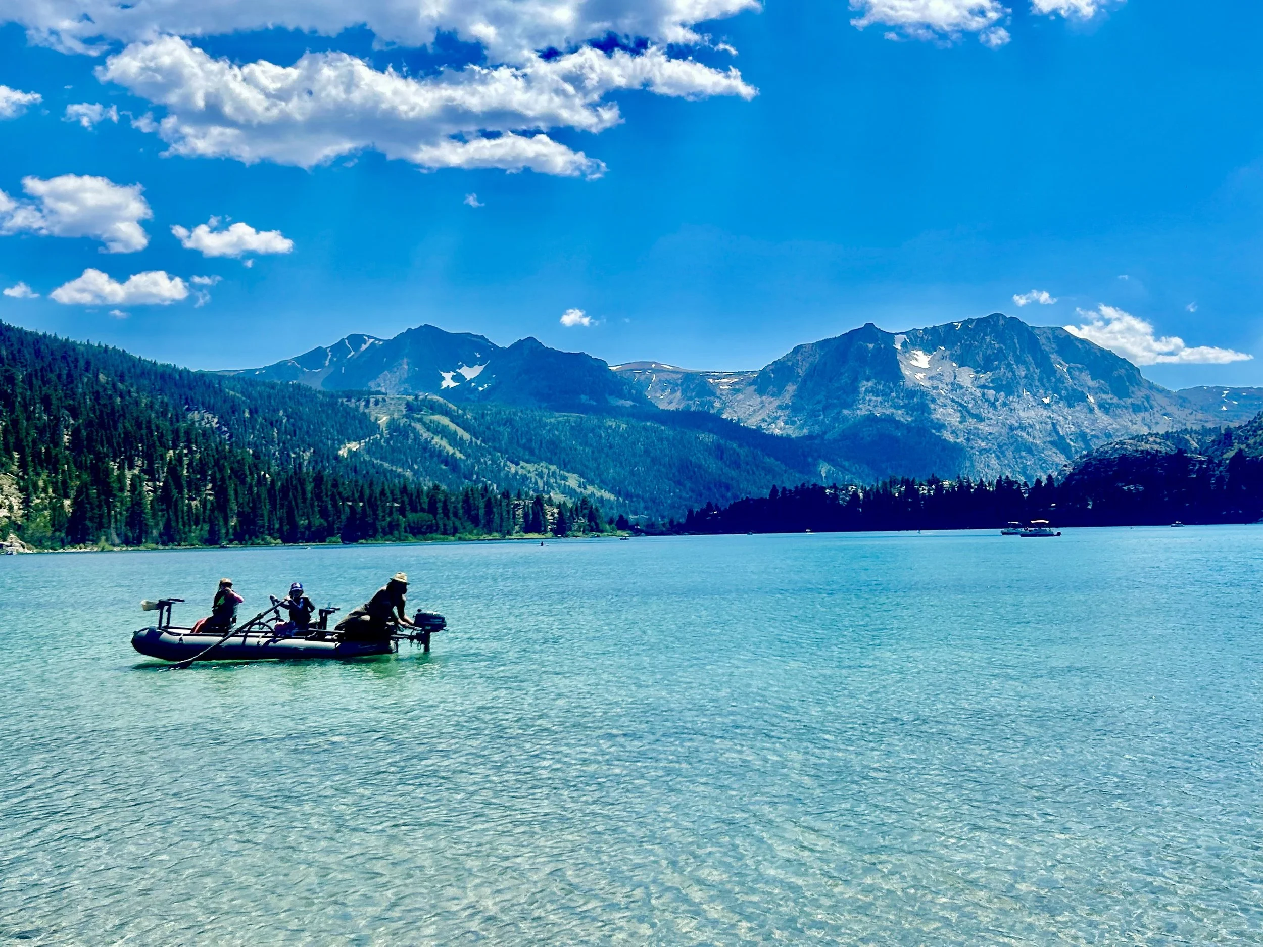 A boat with three people paddling on a turquoise lake surrounded by mountains and a clear blue sky with some clouds.