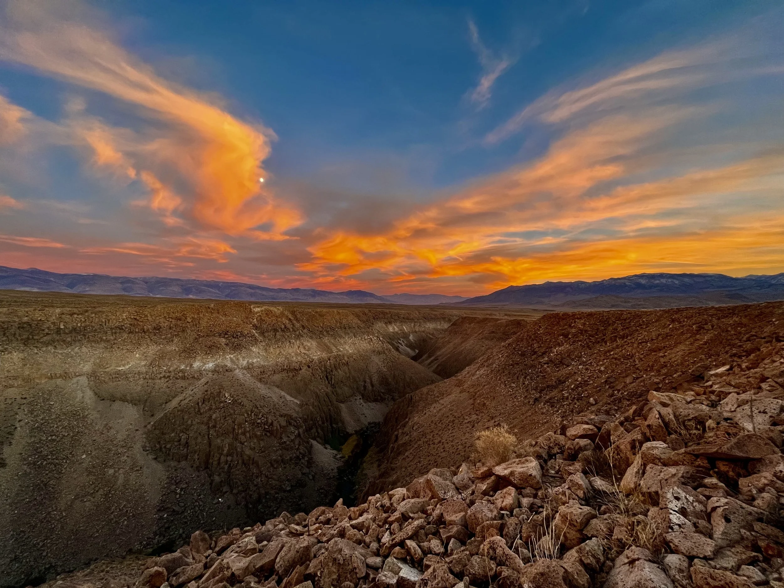 A rugged canyon landscape during sunset with a vibrant orange and pink sky above distant mountains.