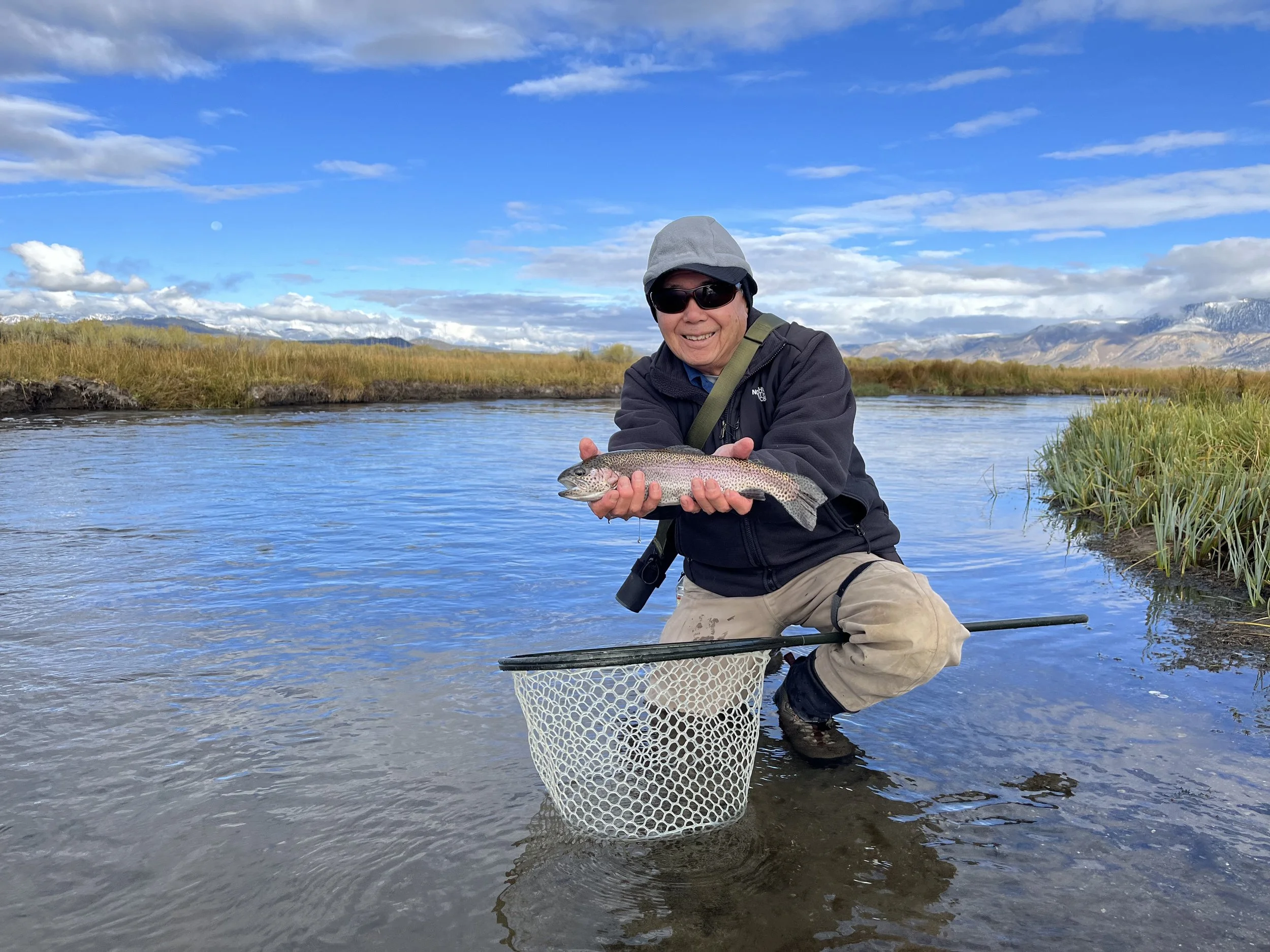Smiling man in outdoor gear kneeling in a river holding a rainbow trout, with a fishing net on the ground nearby, under a blue sky with clouds and mountains in the background.