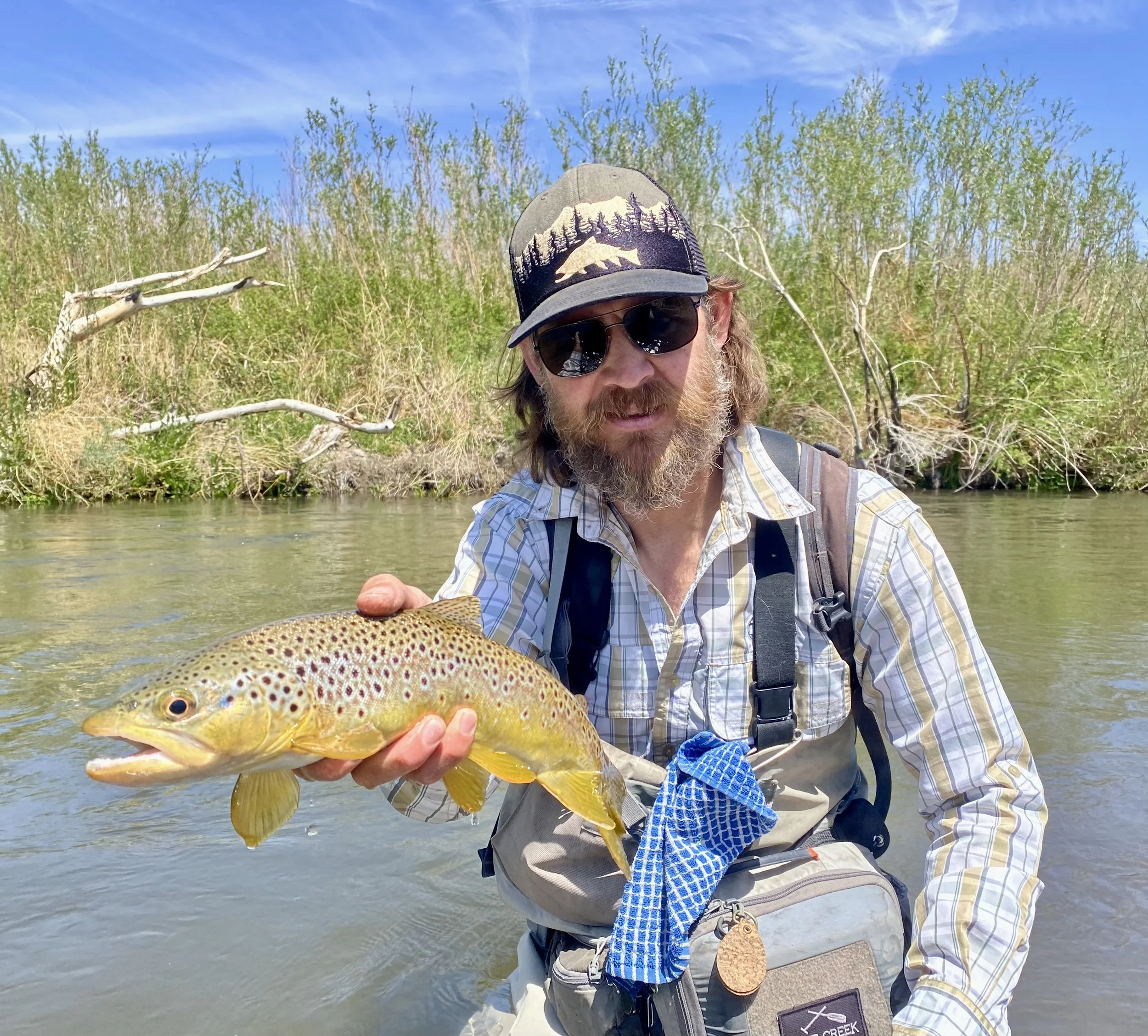 Man in sunglasses wearing a cap and fishing vest holds a rainbow trout in a river with green trees and blue sky in the background.