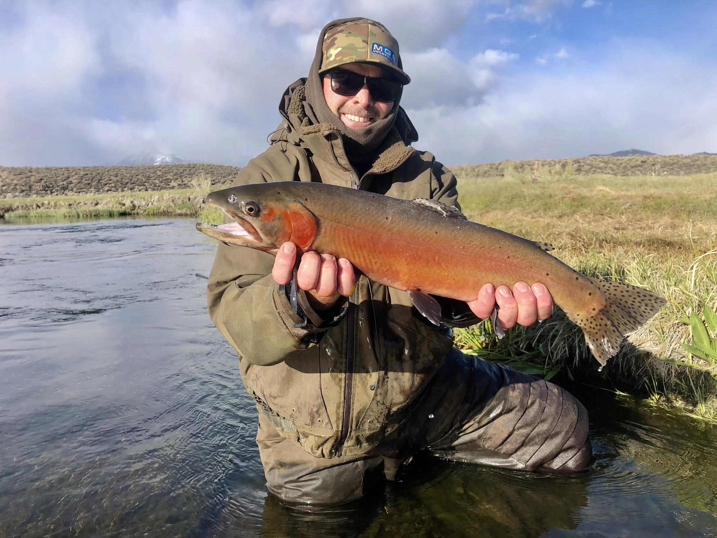 Man wearing camouflage hat, sunglasses, and outdoor gear kneeling in a river, holding a large colorful fish with orange, brown, and greenish hues, with a landscape of grassy hills and cloudy sky in the background.