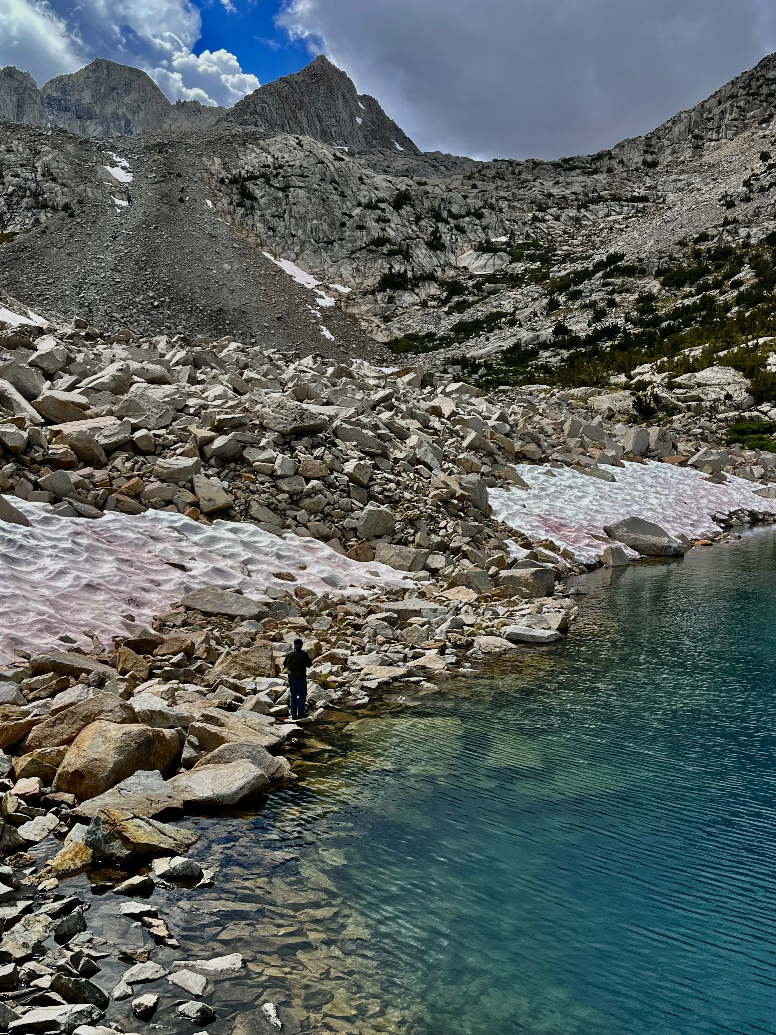 A person standing on rocks at the edge of a mountain lake with rocky, snow patches, rugged mountain terrain, and cloudy sky in the background.