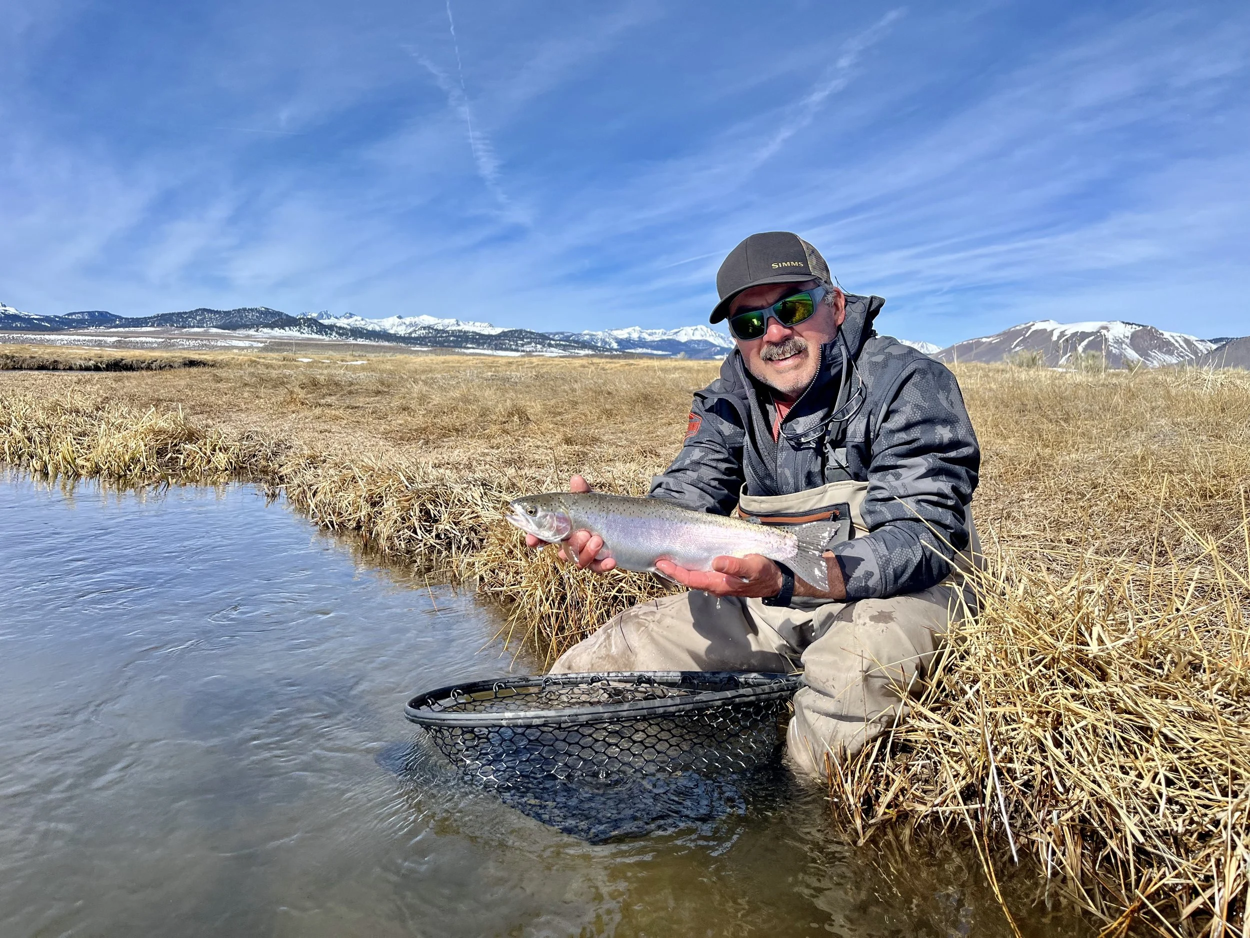 Man wearing sunglasses, a cap, and a waterproof jacket kneeling by a small creek, holding a fish with snow-capped mountains in the background on a clear day.