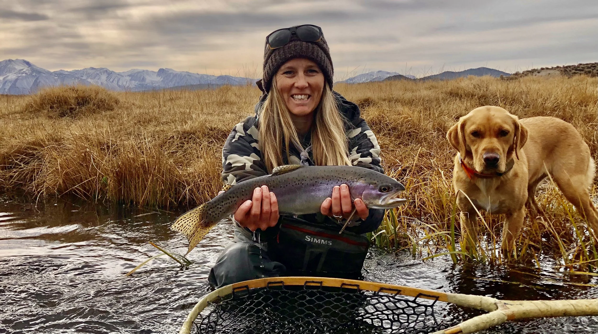 A woman smiling and holding a large fish while kneeling in a river, with a yellow dog nearby and mountains in the background.