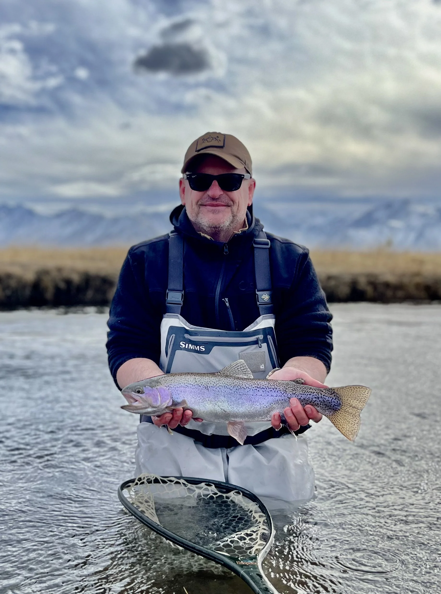 Man wearing sunglasses and a hat holding a rainbow trout in a river with mountains and cloudy sky in the background.