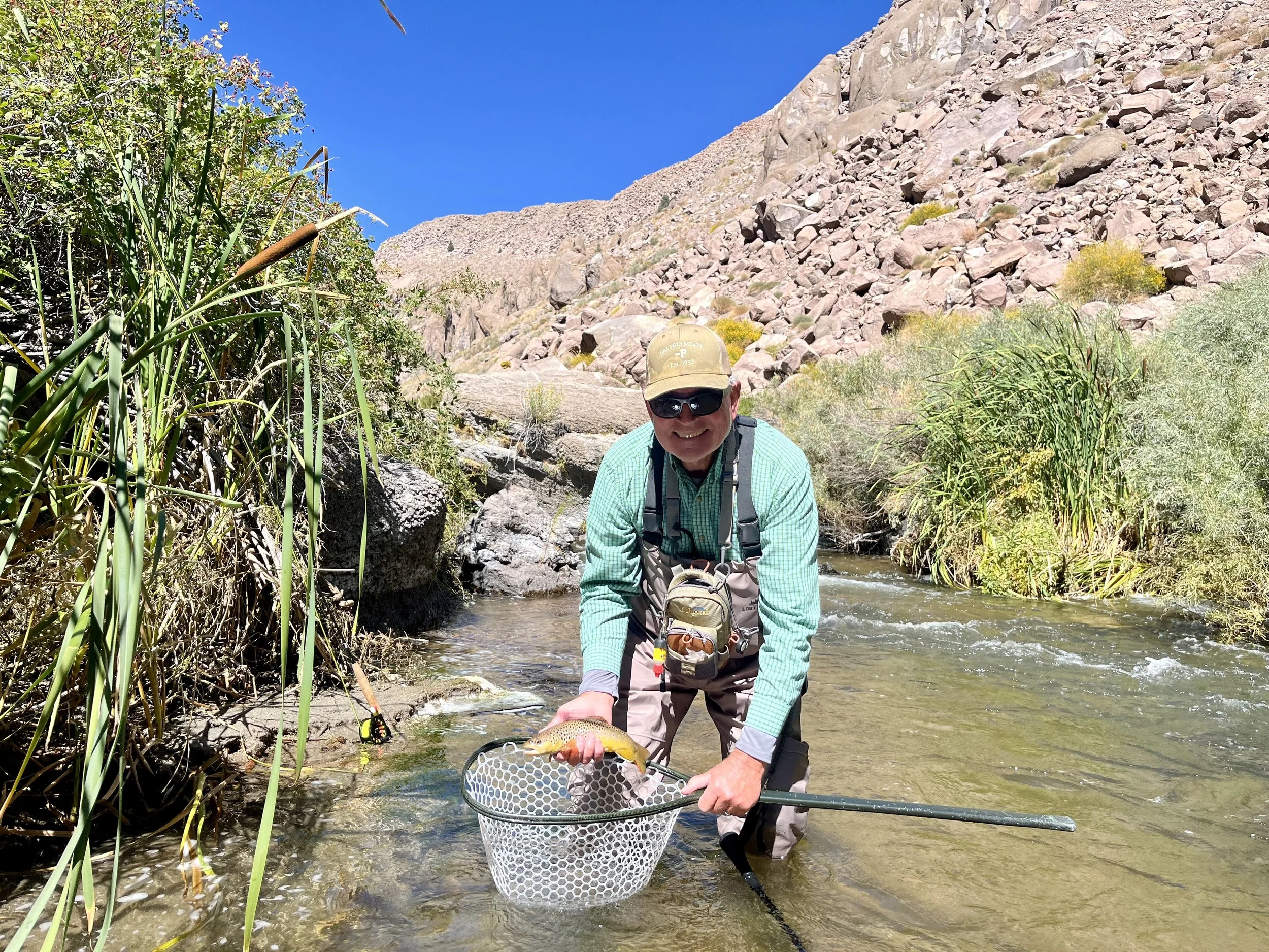 A man wearing sunglasses, a beige hat, and outdoor fishing gear kneeling in a creek holding a fish in a net, surrounded by tall grass and rocky terrain under a clear blue sky.