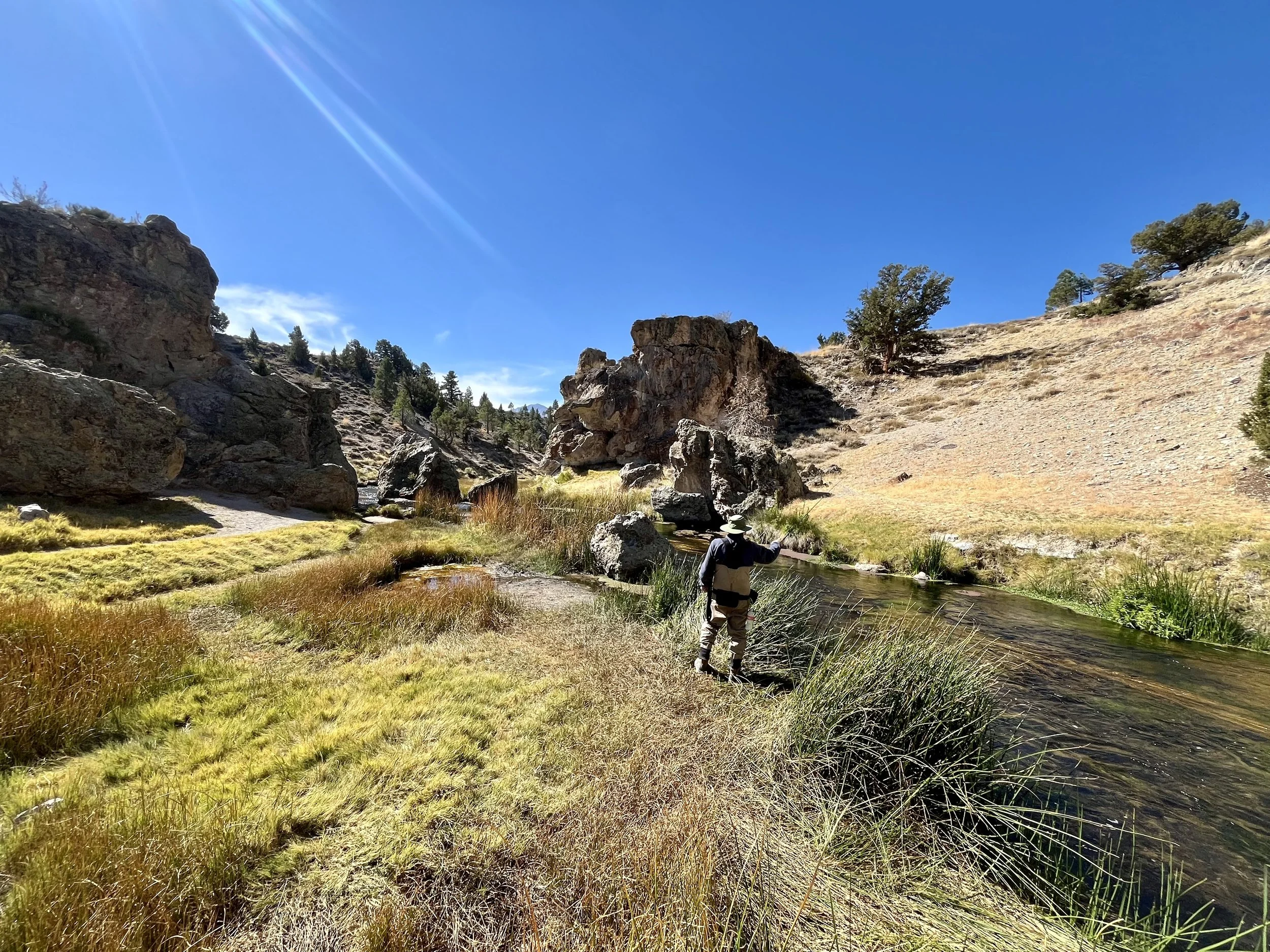 A person standing by a creek in a mountainous landscape under a bright blue sky with the sun shining.