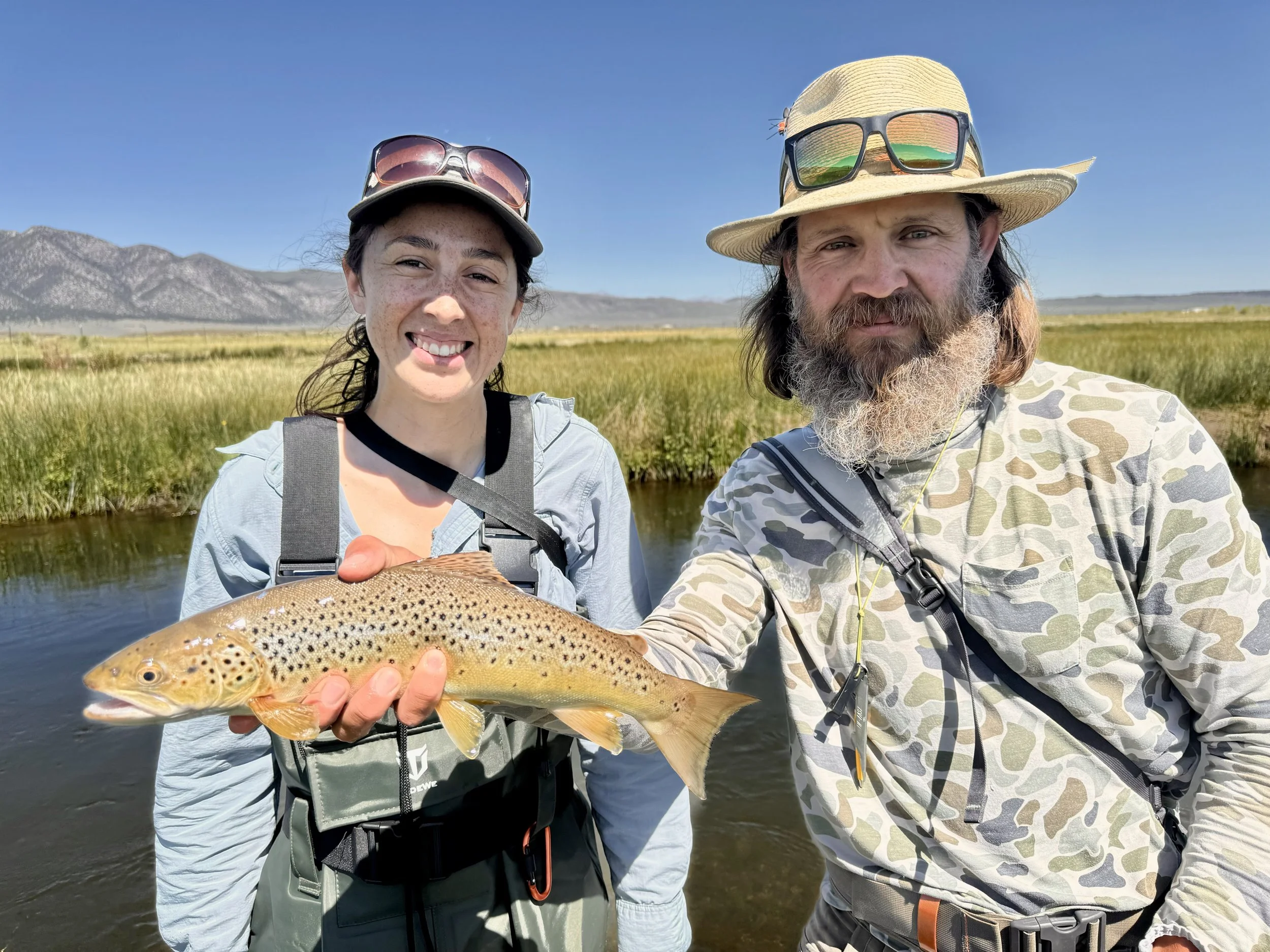 A woman and a man outdoors holding a freshly caught brown trout, with a river, grassy landscape, and mountains in the background on a sunny day.
