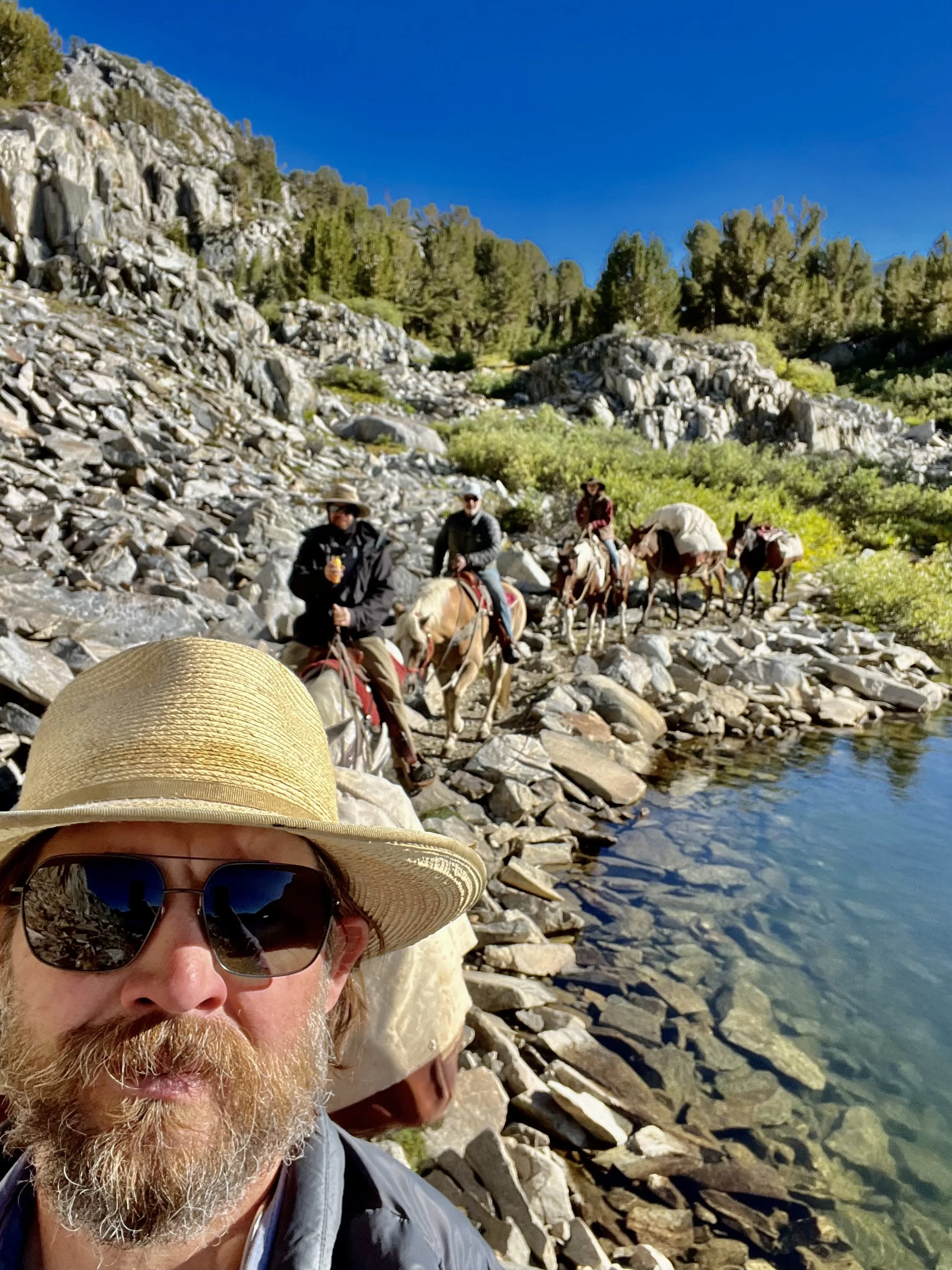 A man with sunglasses and a straw hat taking a selfie by a rocky lakeshore, with three people on horseback in the background along a nature trail in a mountainous area with trees and clear blue sky.