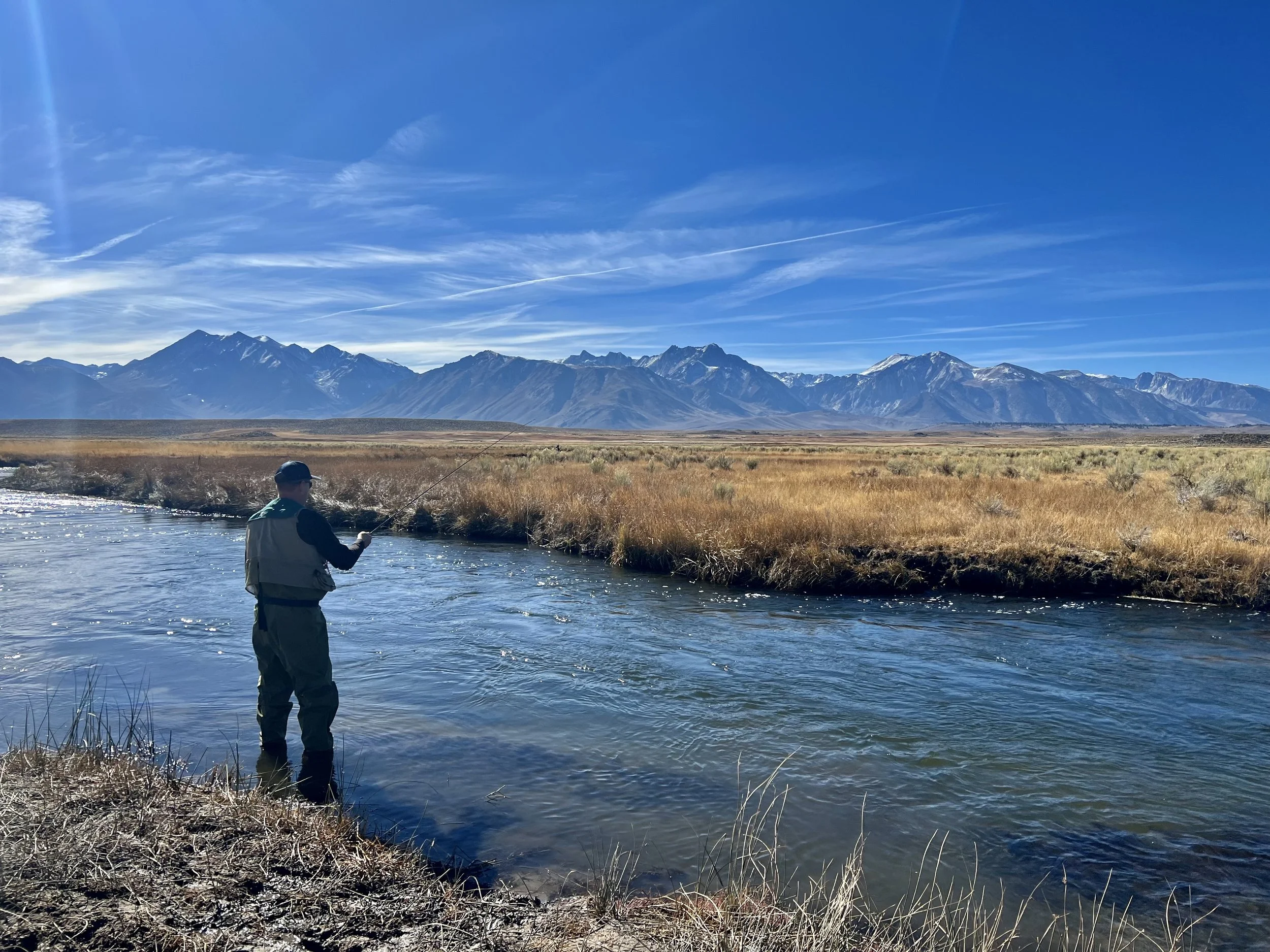 A man fishing in a river with mountains in the background on a sunny day.