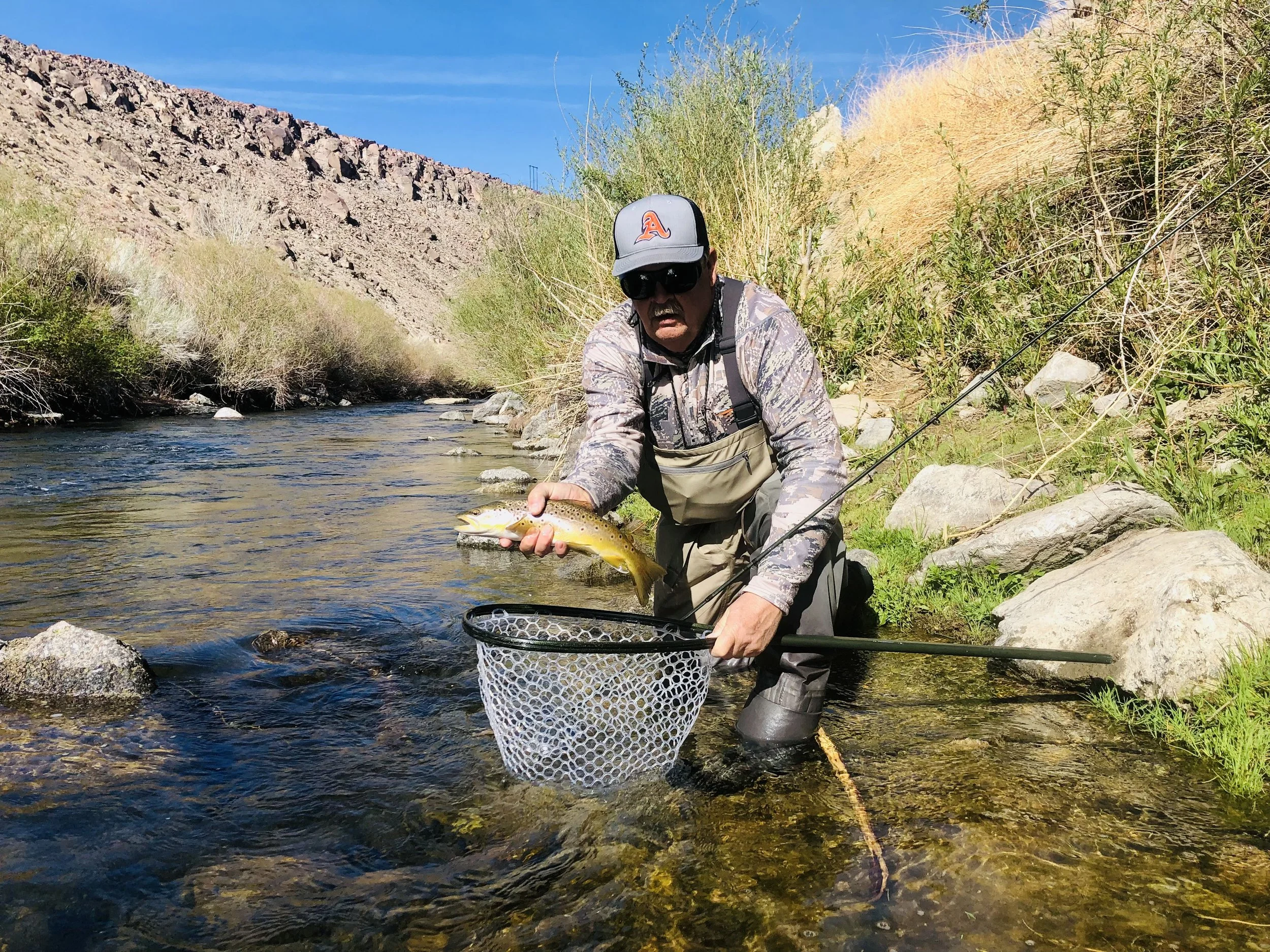 A man in camouflage clothing, sunglasses, and a baseball cap kneels in a shallow river, holding a fish with his right hand and a fishing net with his left, surrounded by rocks and dry vegetation, with mountains and a blue sky in the background.