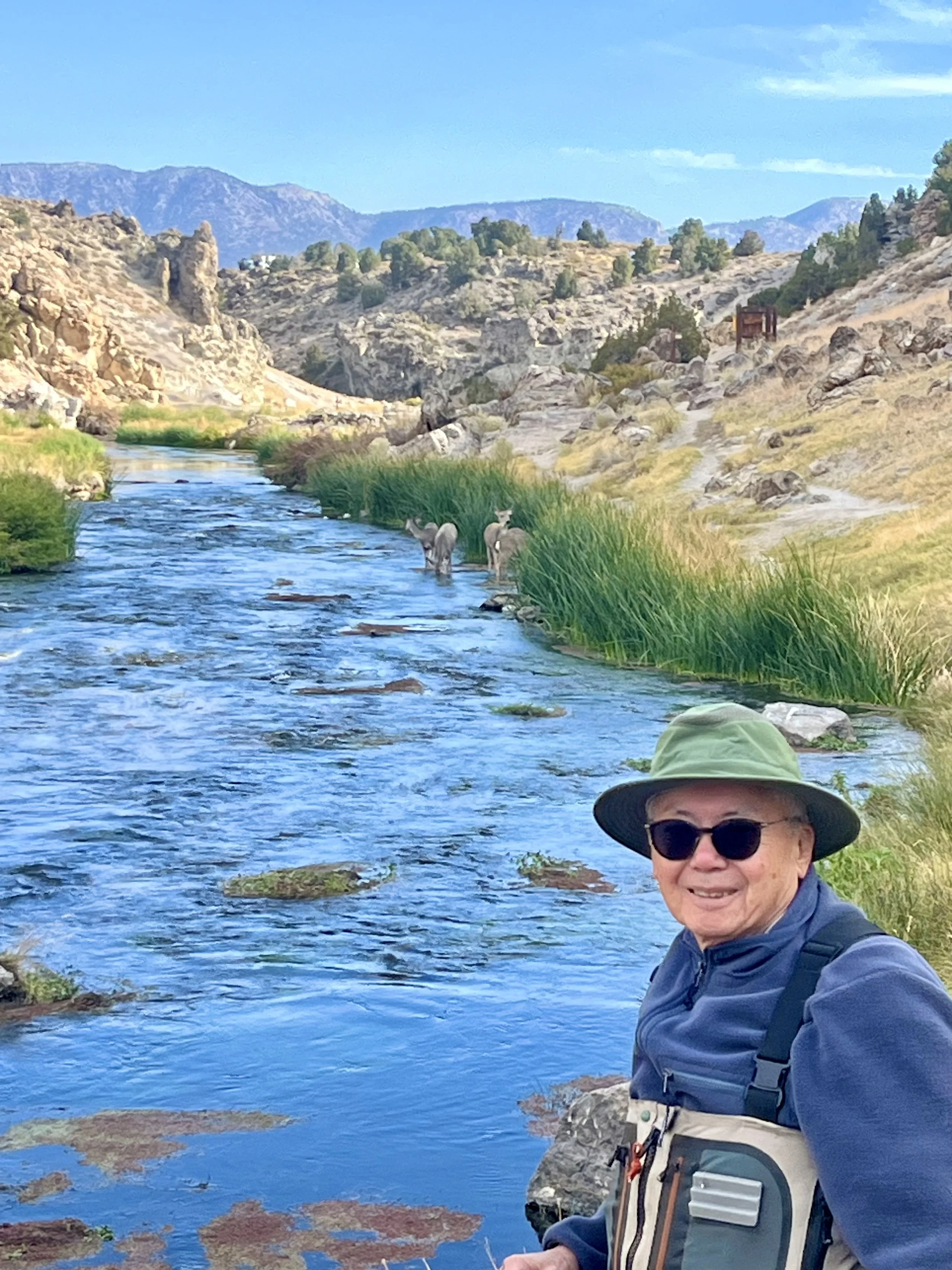 A smiling man in sunglasses and a wide-brimmed hat stands by a stream with two mountain goats in the background, surrounded by rocky hills, tall grass, and distant mountains under a blue sky.