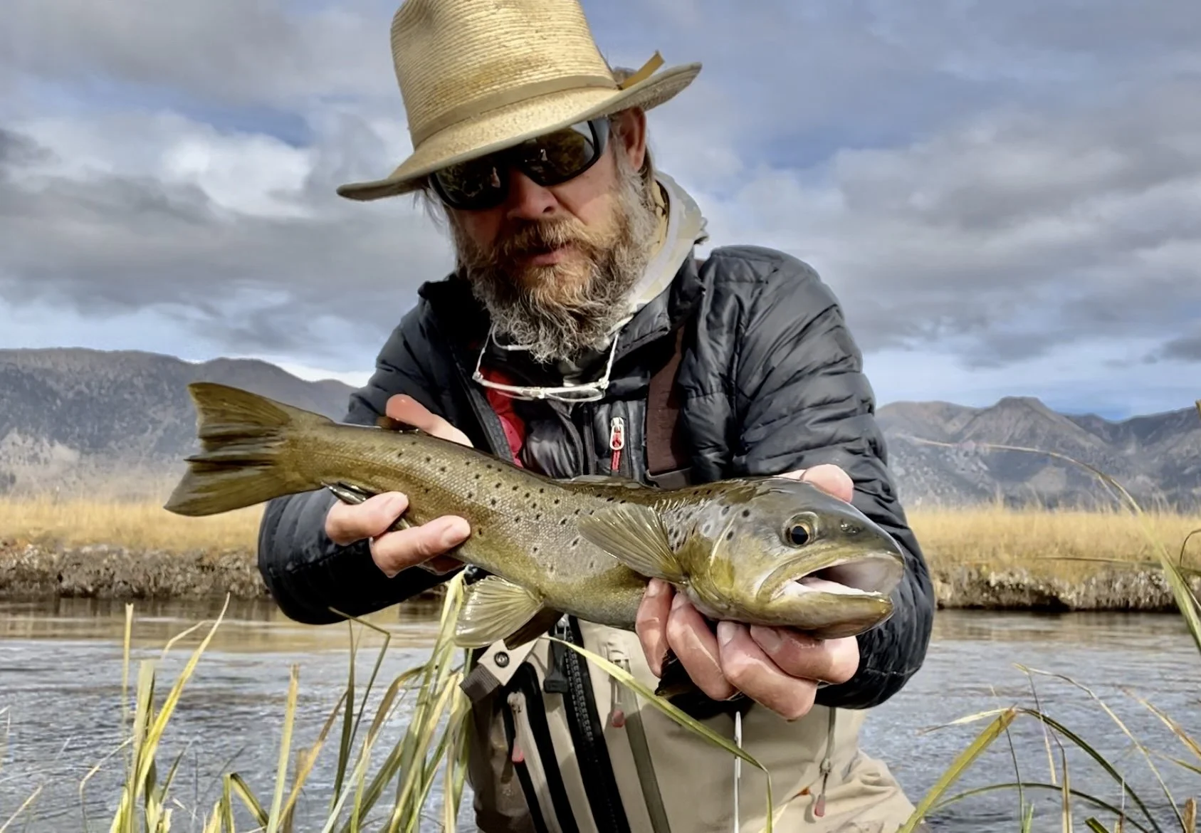 Man in outdoor fishing gear holding a fish he caught near a river, with mountains and cloudy sky in the background.