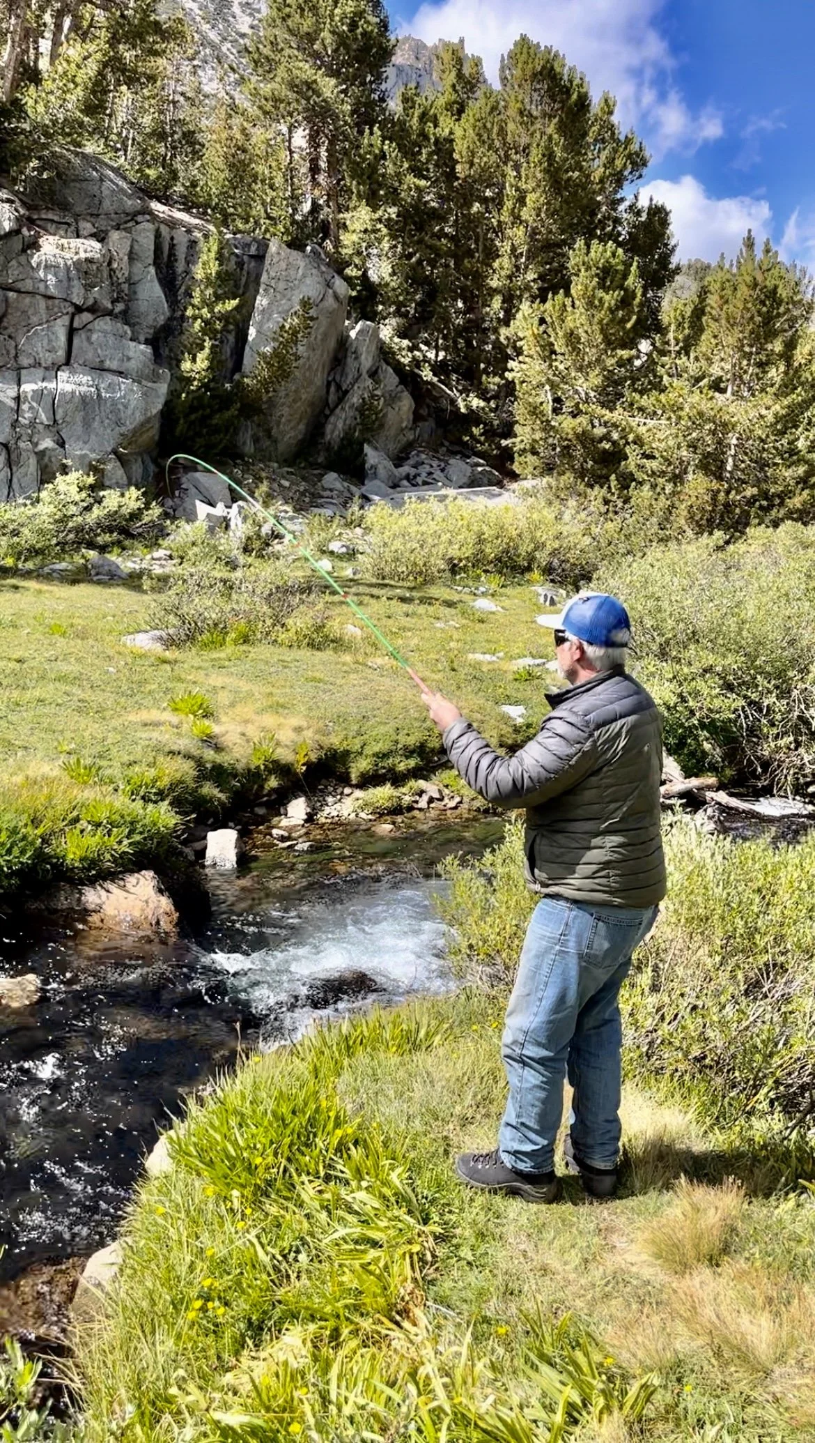 A man stands on grassy bank fishing in a river surrounded by pine trees and rocky cliffs under a partly cloudy sky.