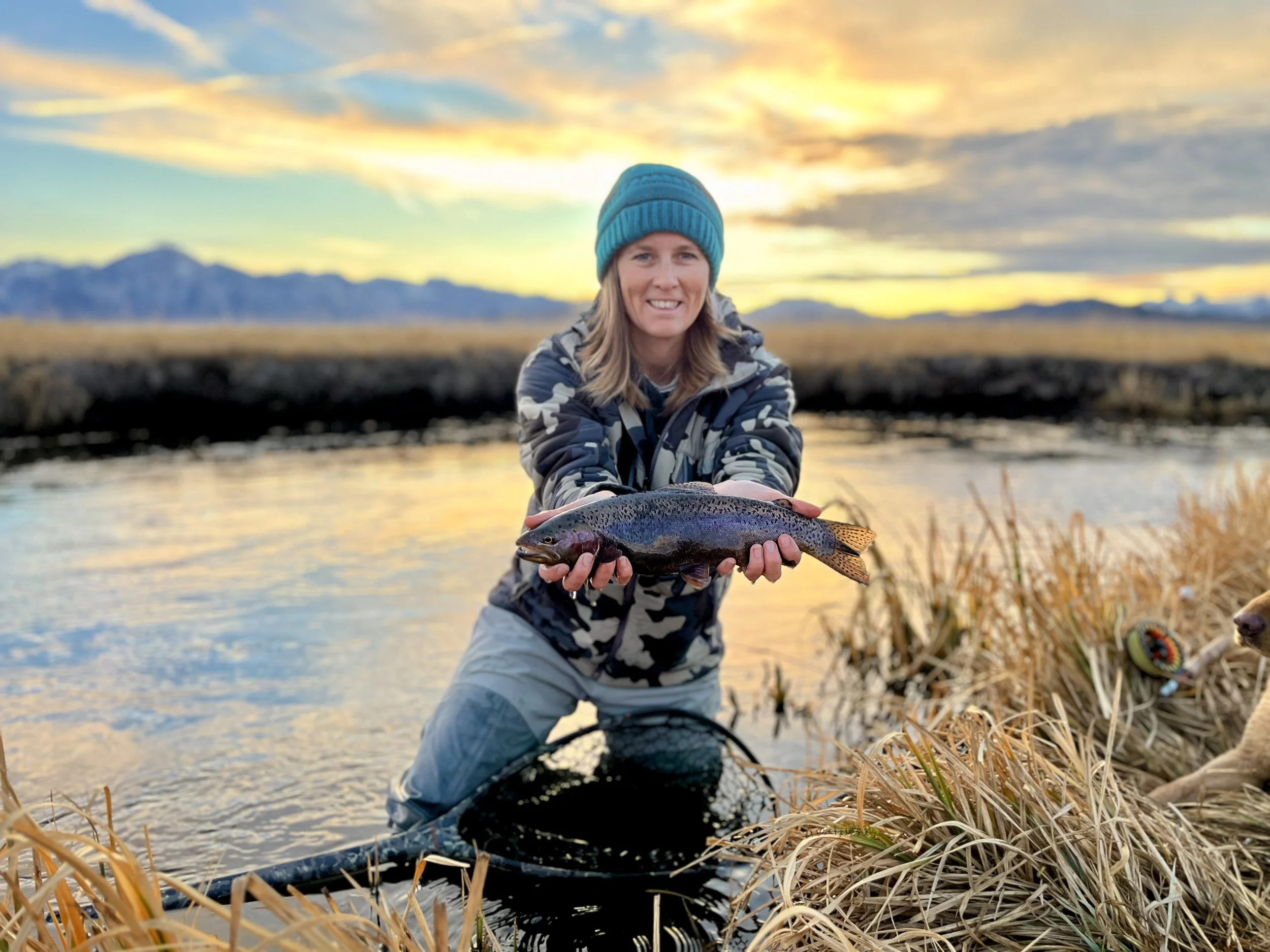A woman in outdoor gear holding a large fish by a river at sunset.