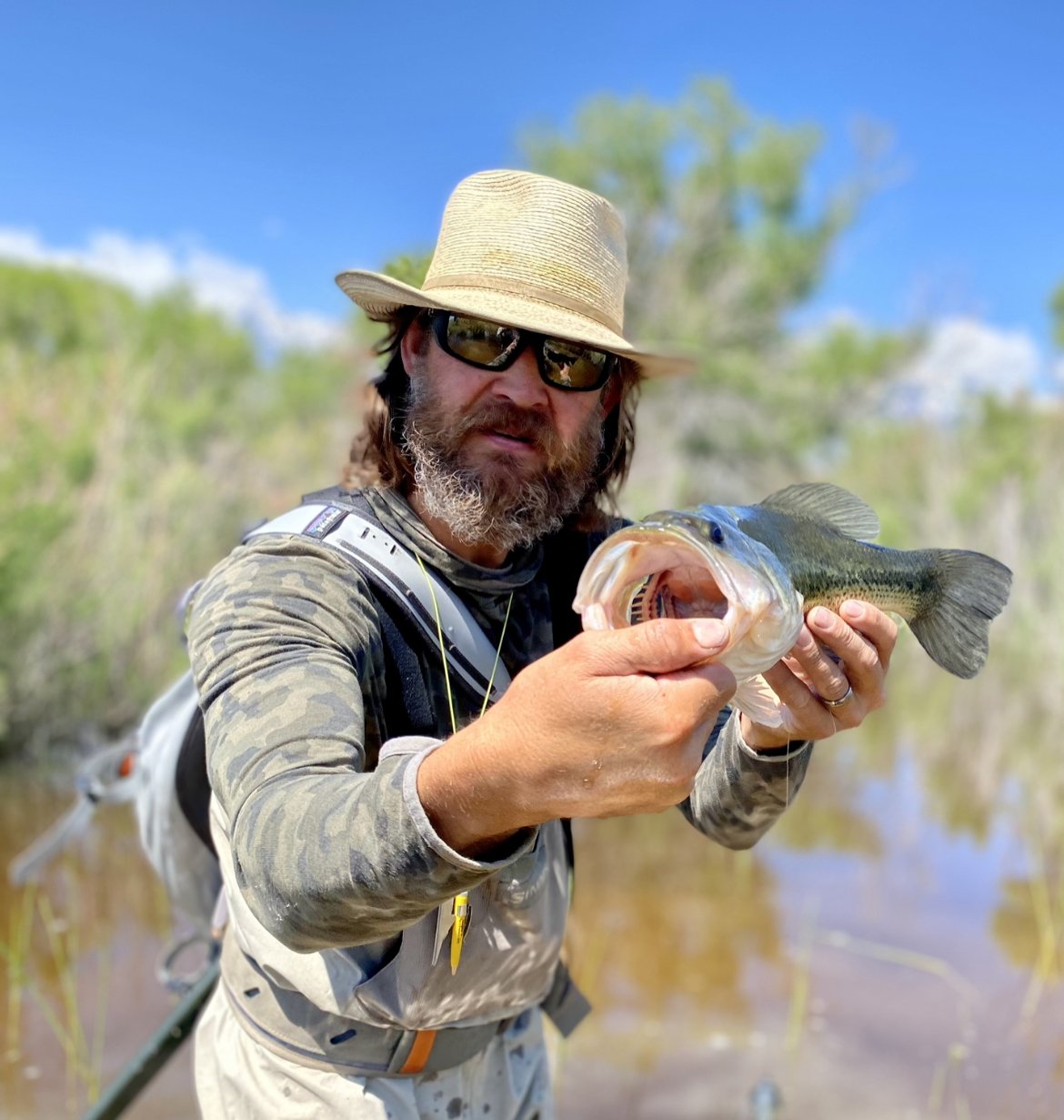 Man wearing a straw hat, sunglasses, and camouflage shirt holding a largemouth bass fish outdoors near a pond or lake with trees and blue sky in the background.