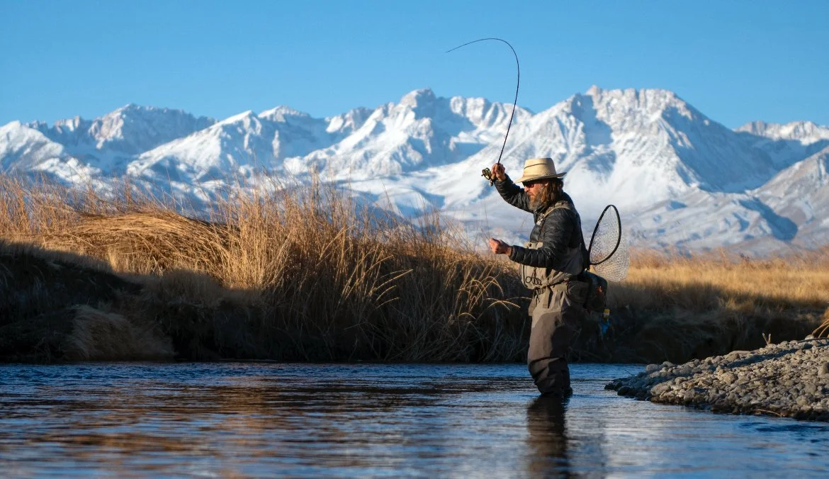 A person fishing in a river with snow-capped mountains in the background.