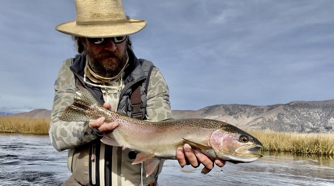 Man wearing a straw hat and camouflage jacket holding a large fish in a river with grassy banks and mountains in the background.