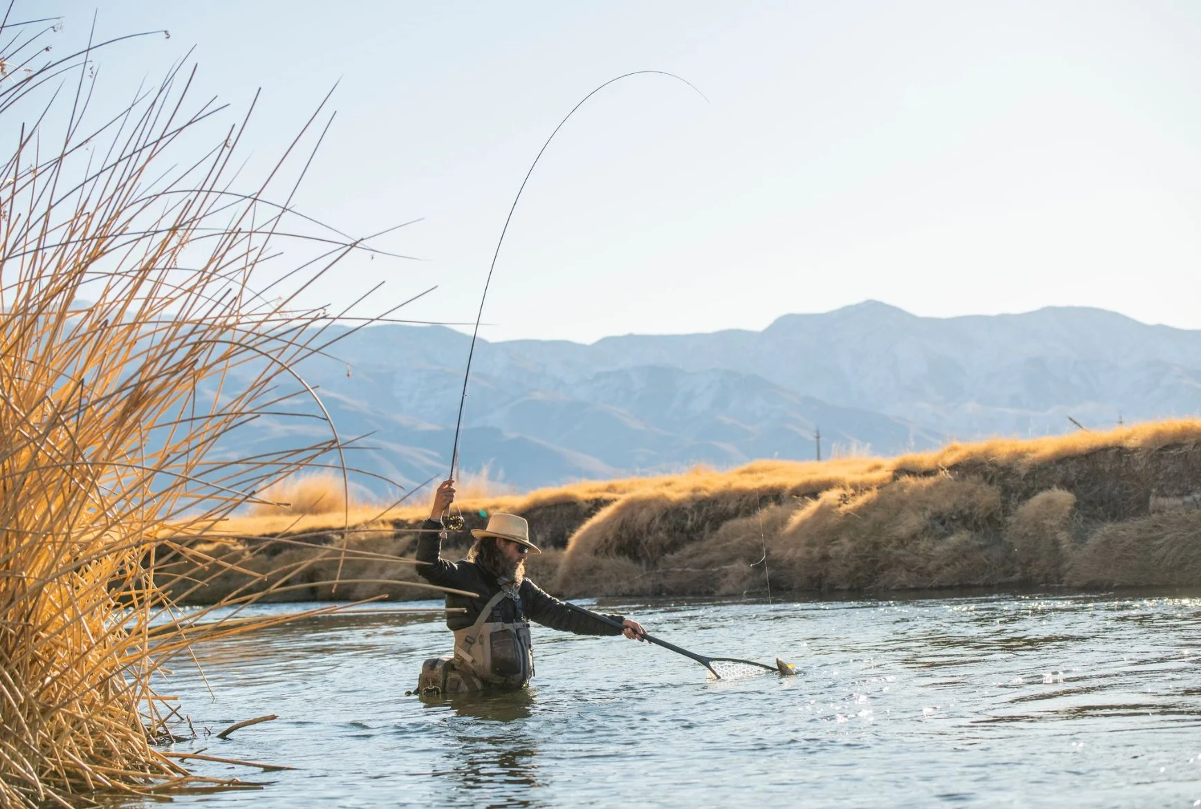 Person fishing in a river with mountains in the background and dry grass along the riverbank.